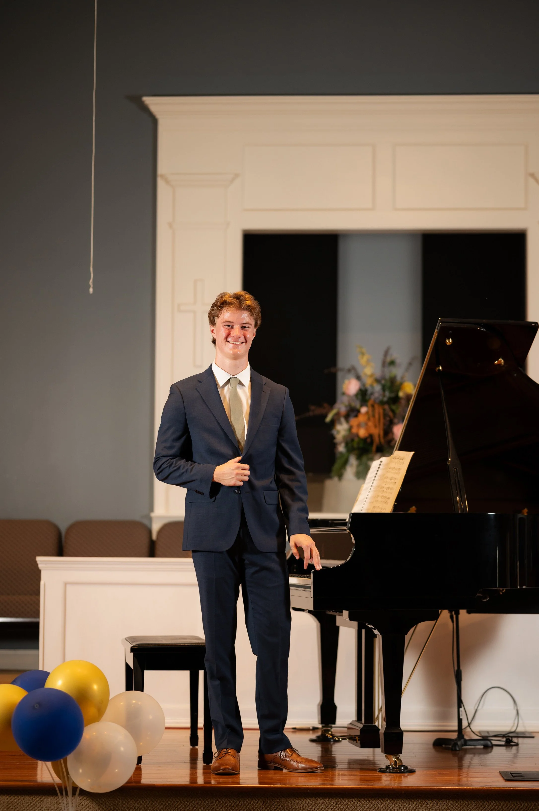 A young man in a suit standing next to a grand piano in a church or auditorium setting, smiling at the camera.