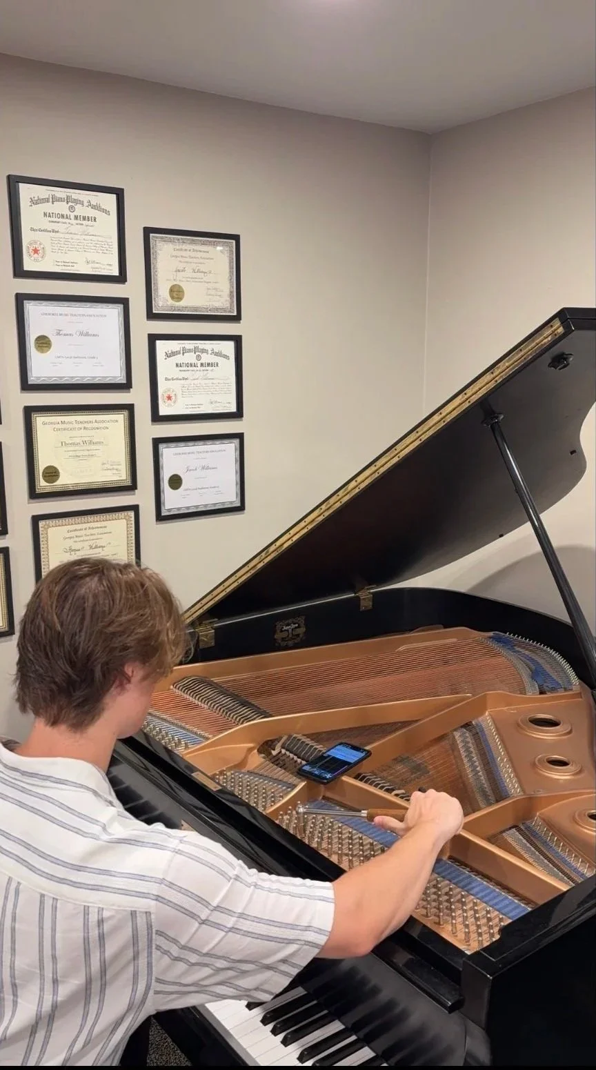 Person playing a grand piano with a mobile phone resting on the strings, wall with framed certificates or awards above the piano.
