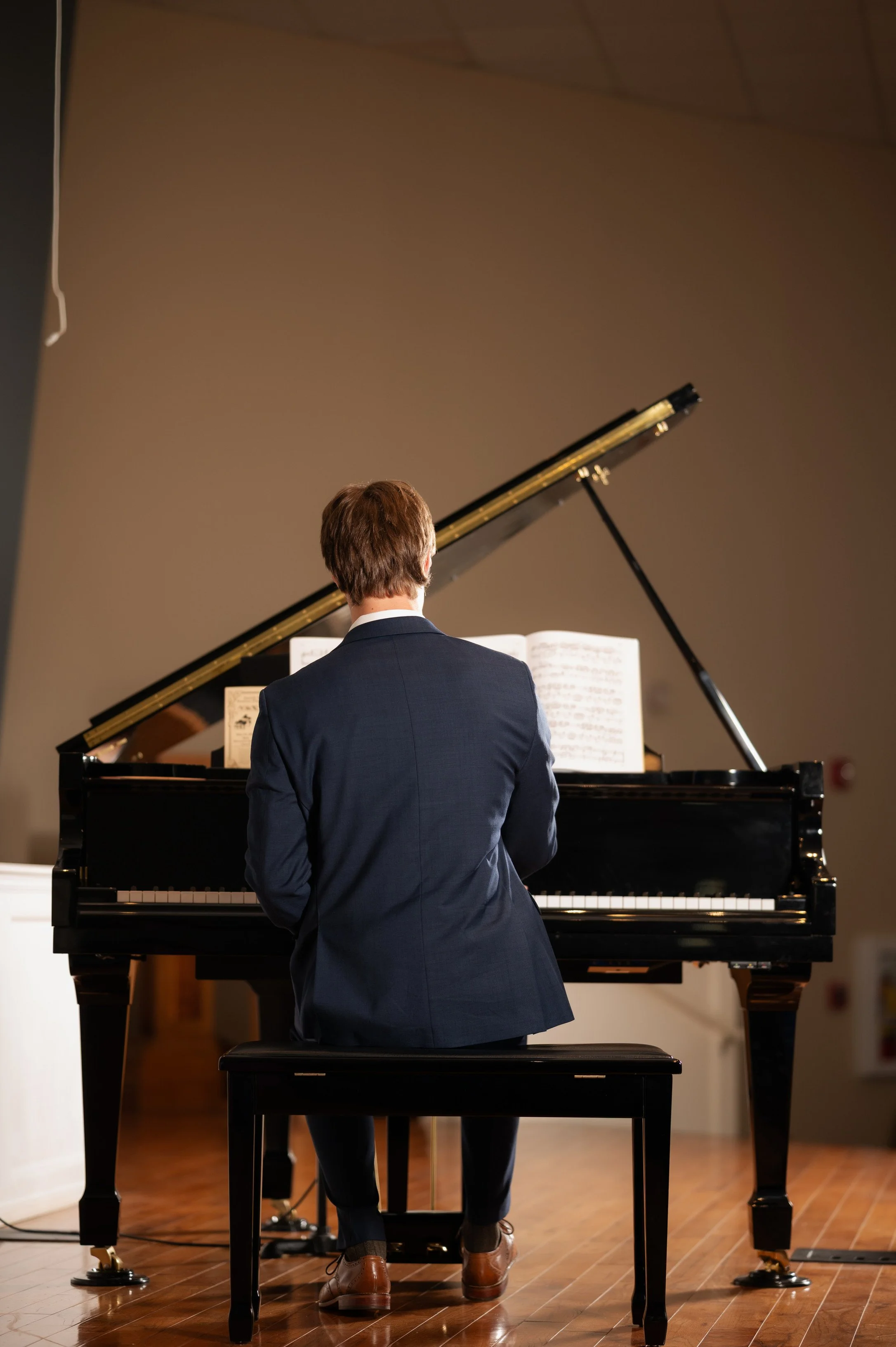 A man in a navy suit and brown shoes playing a grand piano indoors, seen from behind, with sheet music on the piano, against a plain brown wall.