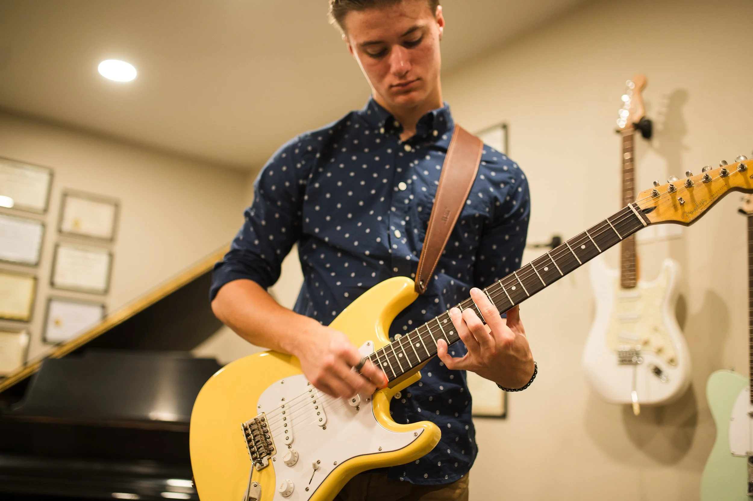 A young man playing a yellow Fender electric guitar indoors, with guitars on the wall behind him.