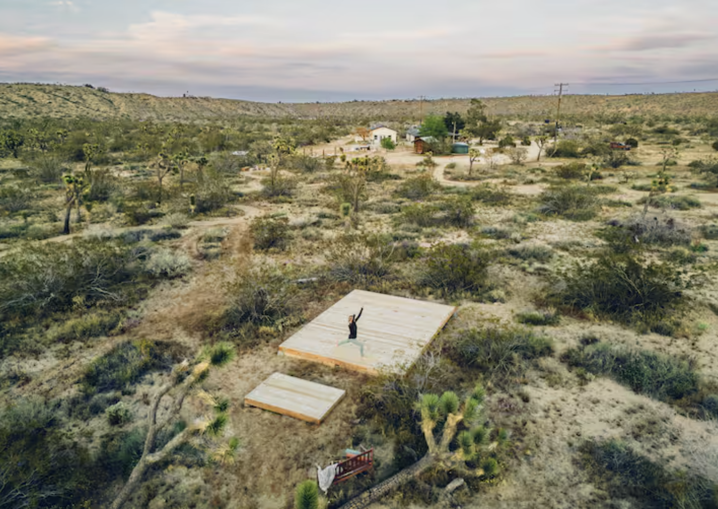A person standing on a wooden platform in a desert landscape with sparse vegetation, surrounding small bushes and cacti, with a few houses and power lines in the background under a cloudy sky.