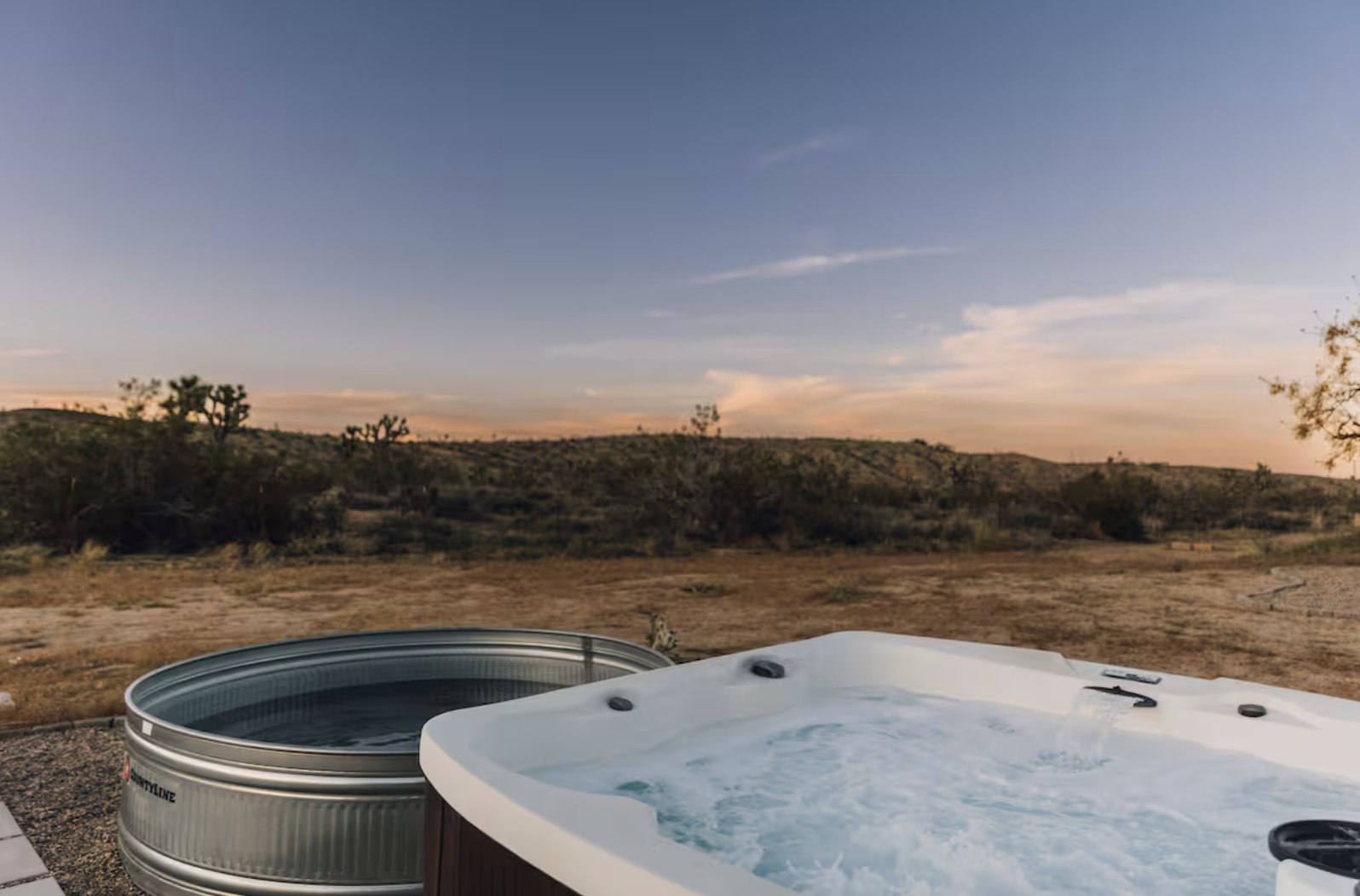 An outdoor hot tub filled with water and jets, next to a fire pit in a desert landscape during sunset, with distant hills and sparse vegetation.
