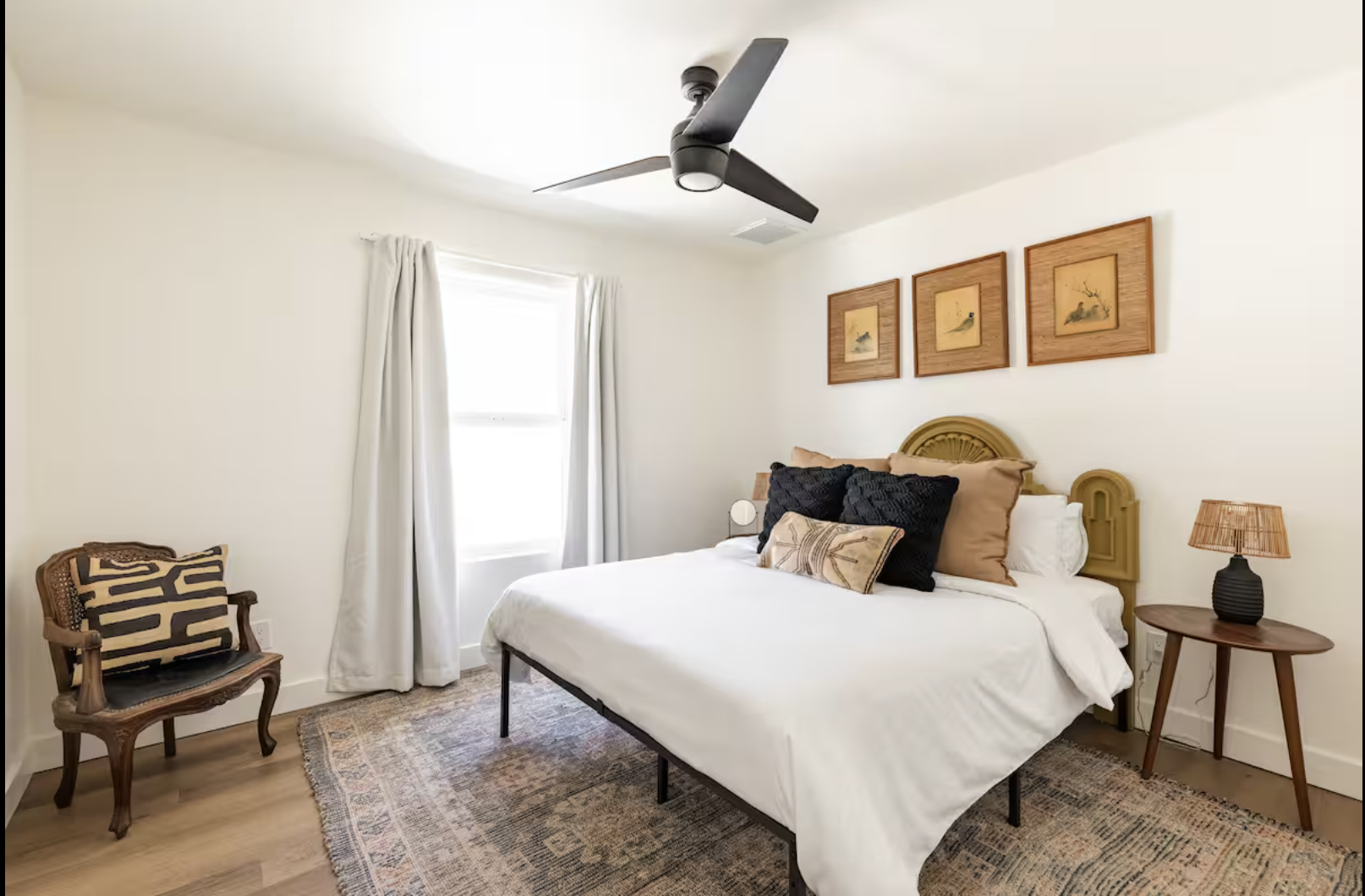A bedroom with a white bed, black and beige pillows, a wooden bedside table with a lamp, a wooden chair with a patterned cushion, white curtains, a window, a ceiling fan, and framed bird artwork on the wall.