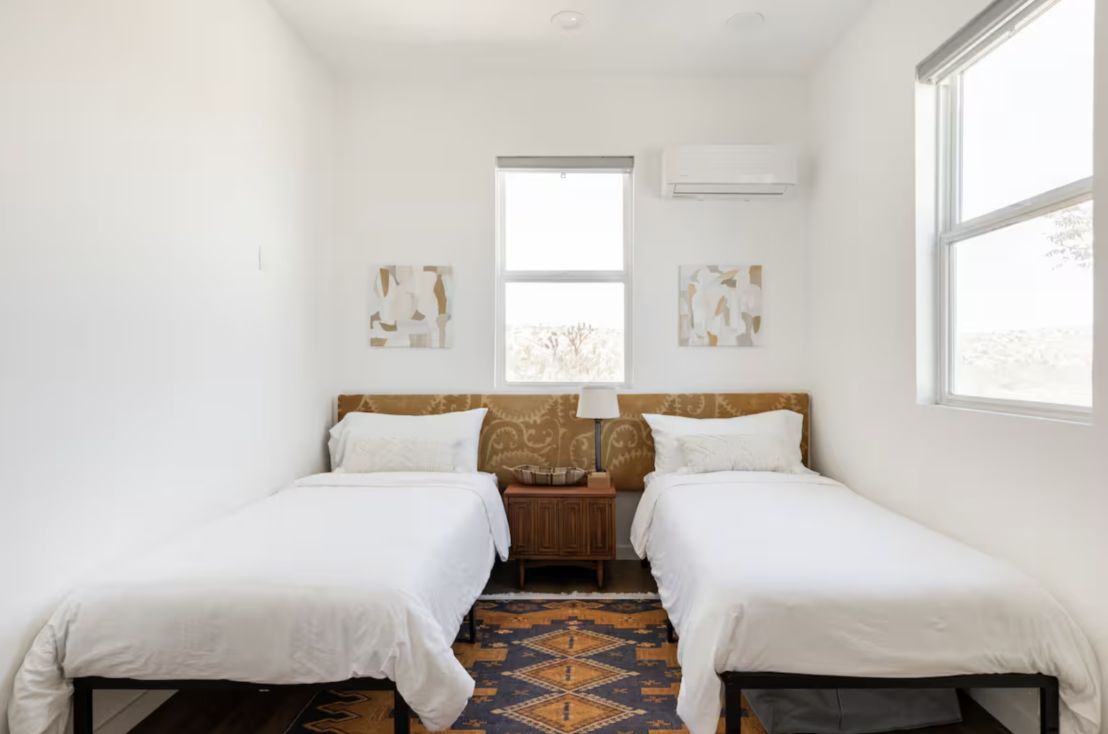 A bedroom with white walls, two beds with white bedding and beige pillows, a wooden nightstand with a lamp and a basket, an air conditioning unit above, two abstract art pieces on the wall, and windows letting in natural light.