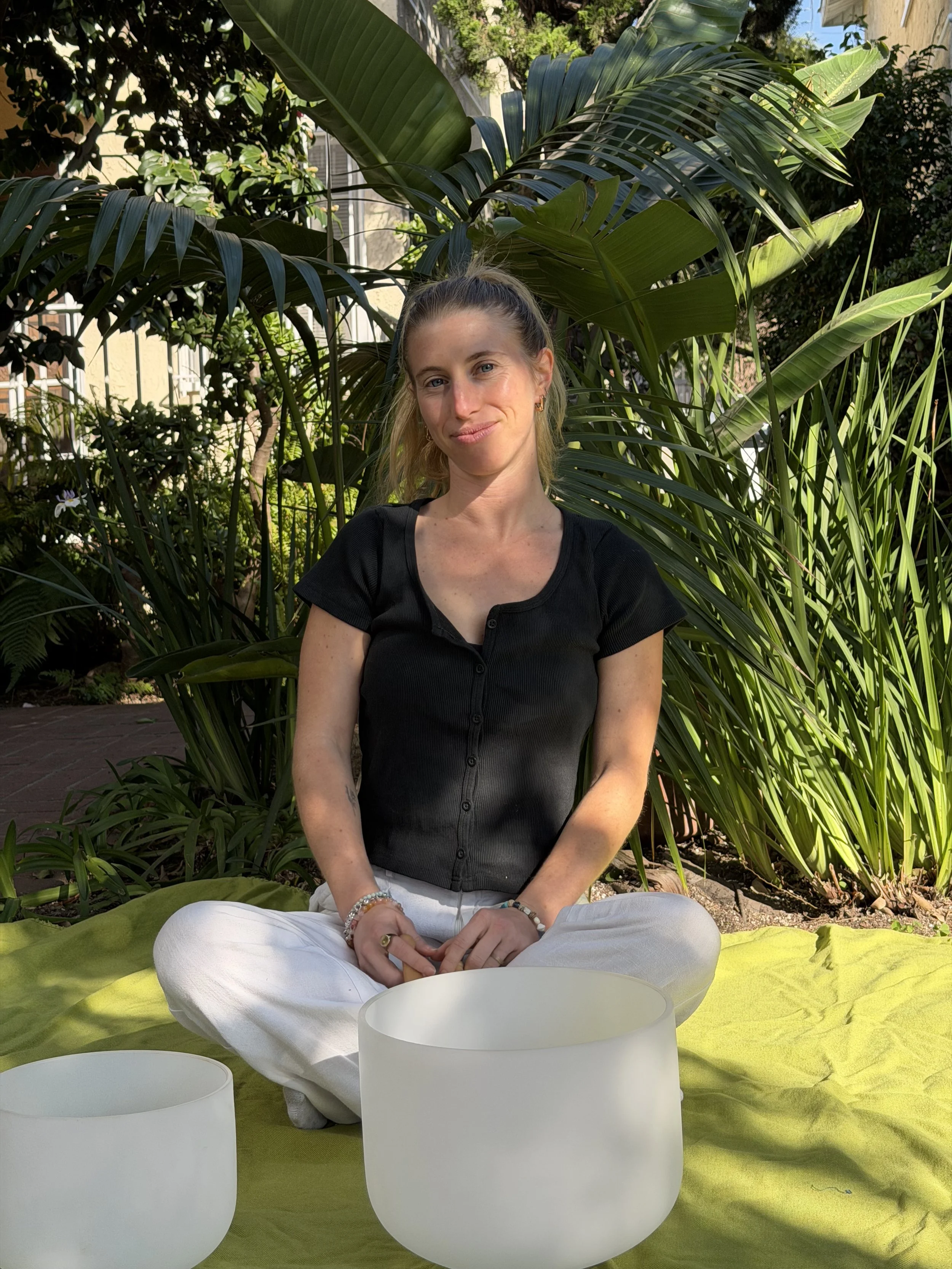 A woman sitting cross-legged on a yellow blanket outdoors, surrounded by green tropical plants, wearing a black top and white pants, with three white sound bowls in front of her.