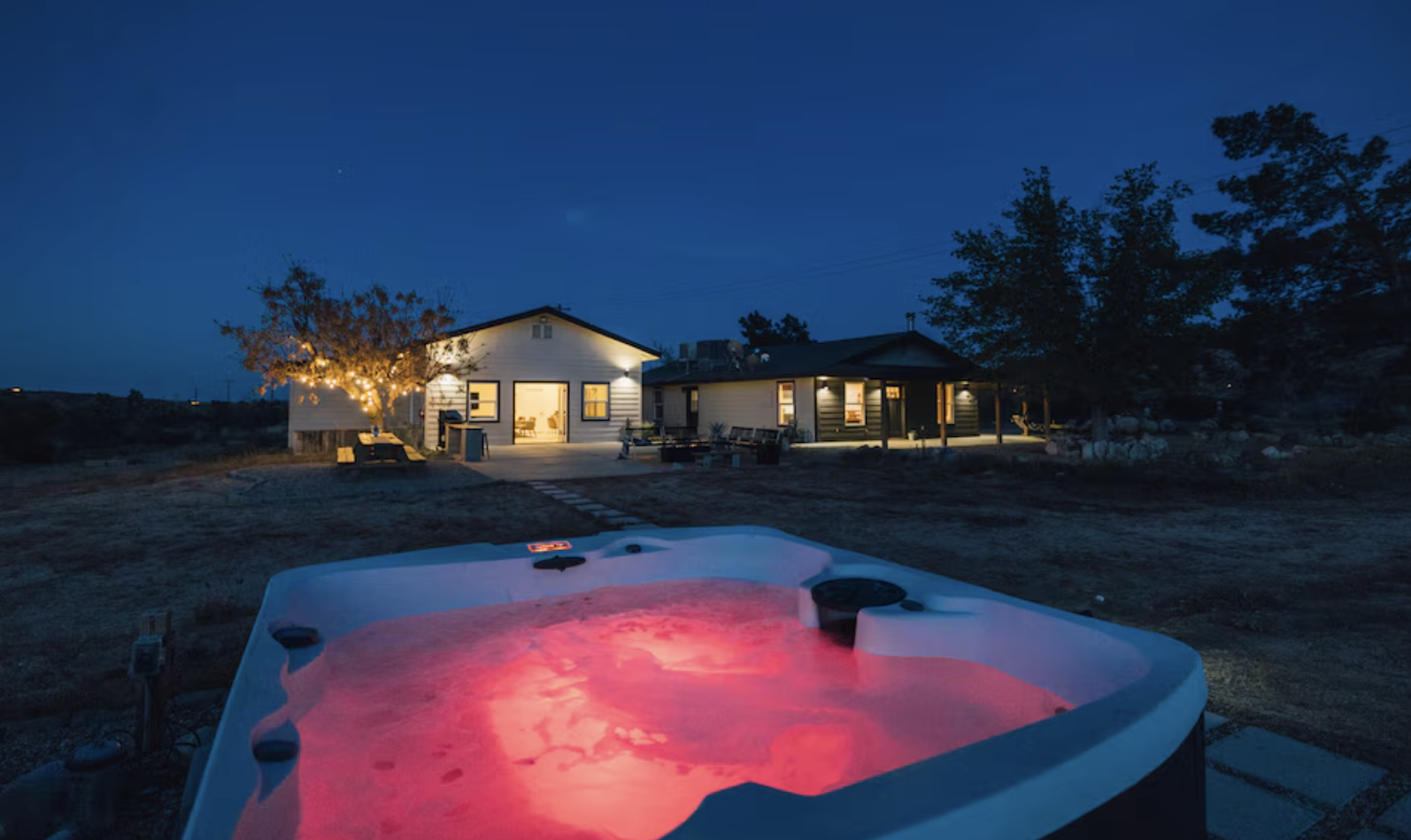 Nighttime outside view of a house with lit windows and outdoor seating area, with a hot tub filled with water glowing red in the foreground.