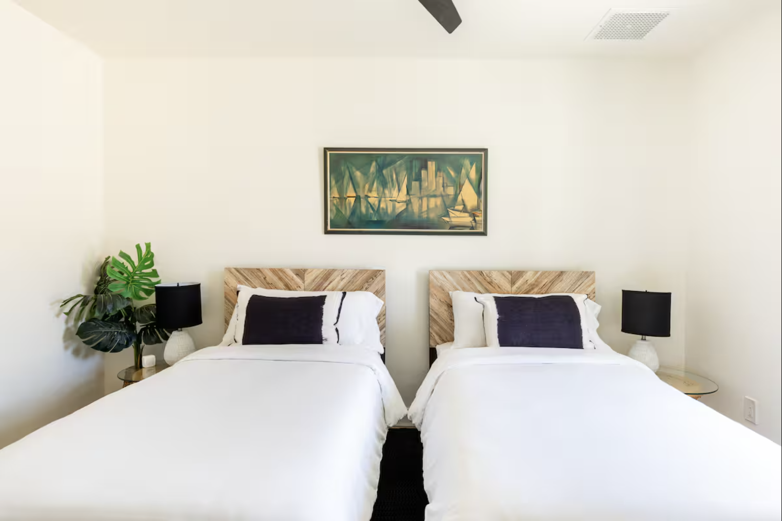 A bedroom with two beds featuring wooden headboards, black pillows, white bedding, and black lamps on transparent side tables. There is a large green plant on the left and a framed abstract painting of a cityscape above the beds on the white wall.