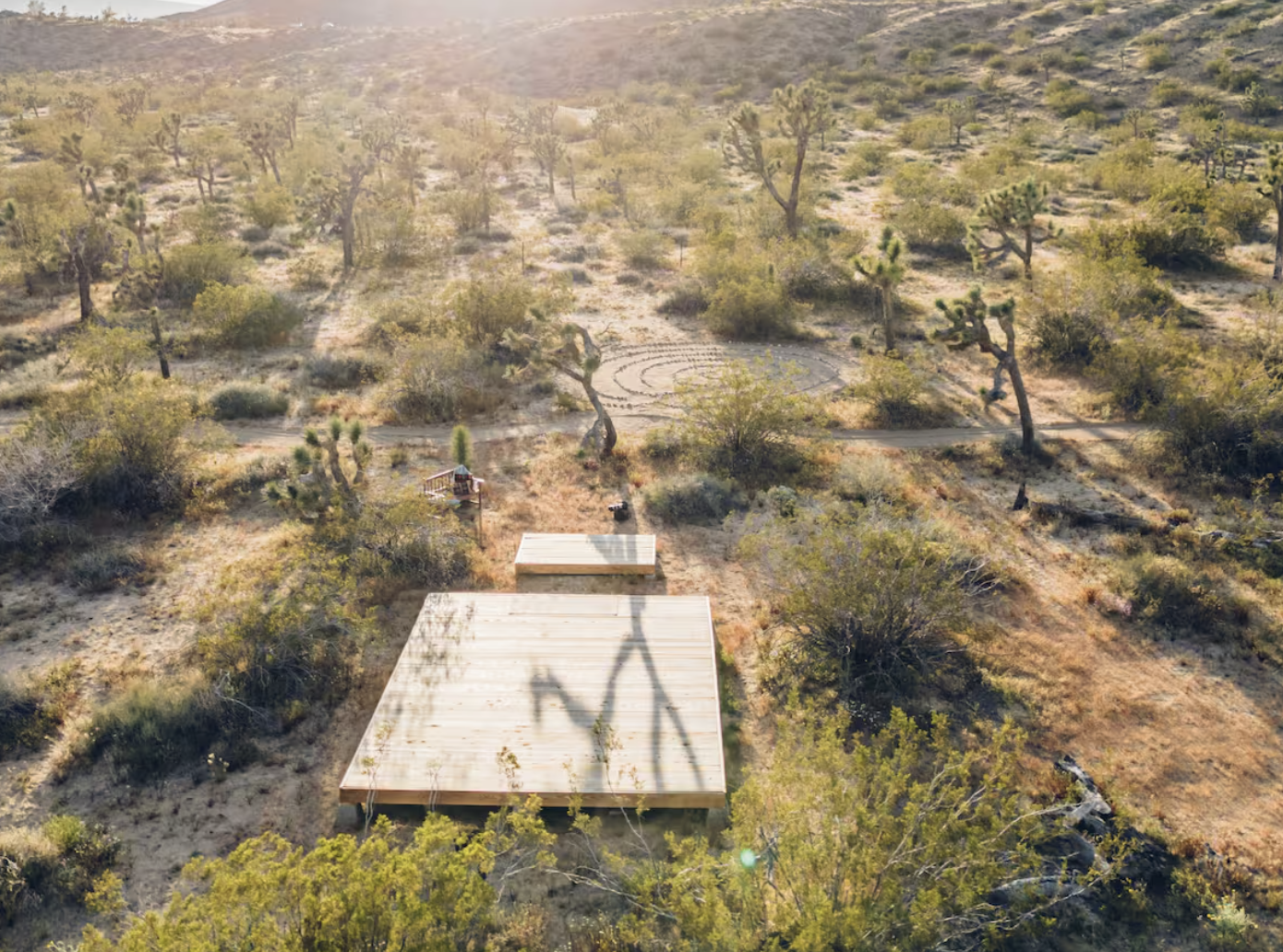 Aerial view of a desert landscape with sparse trees and bushes, a wooden deck with a shadow of a utility pole, and a spiral maze or pattern made in the ground with rocks or dirt.