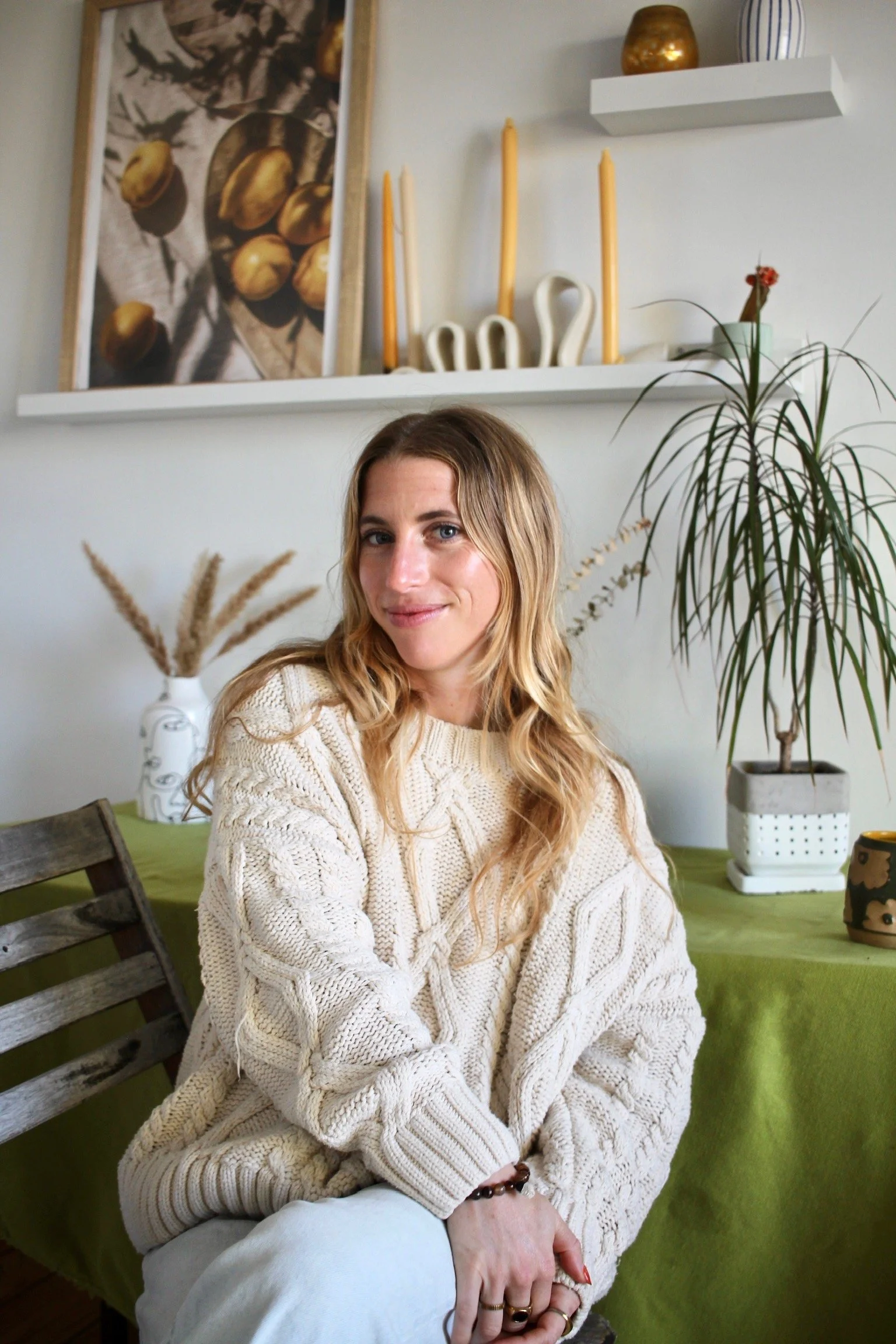 A woman with long blonde hair sitting indoors on a green-covered table in front of a white wall with decorative items, including candles, vases, and artwork. She is wearing a cream-colored knitted sweater and has rings and a bracelet on her left hand.