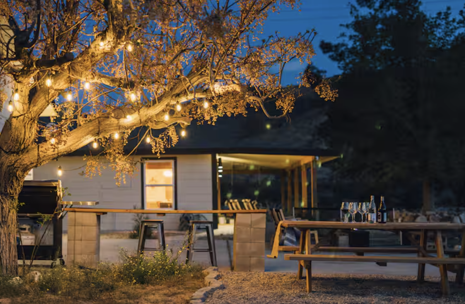 An outdoor evening scene featuring a large tree decorated with string lights, adjacent to a modern house with a porch. There is a picnic table with bottles and glasses, and a bar-height table with stools below the tree.