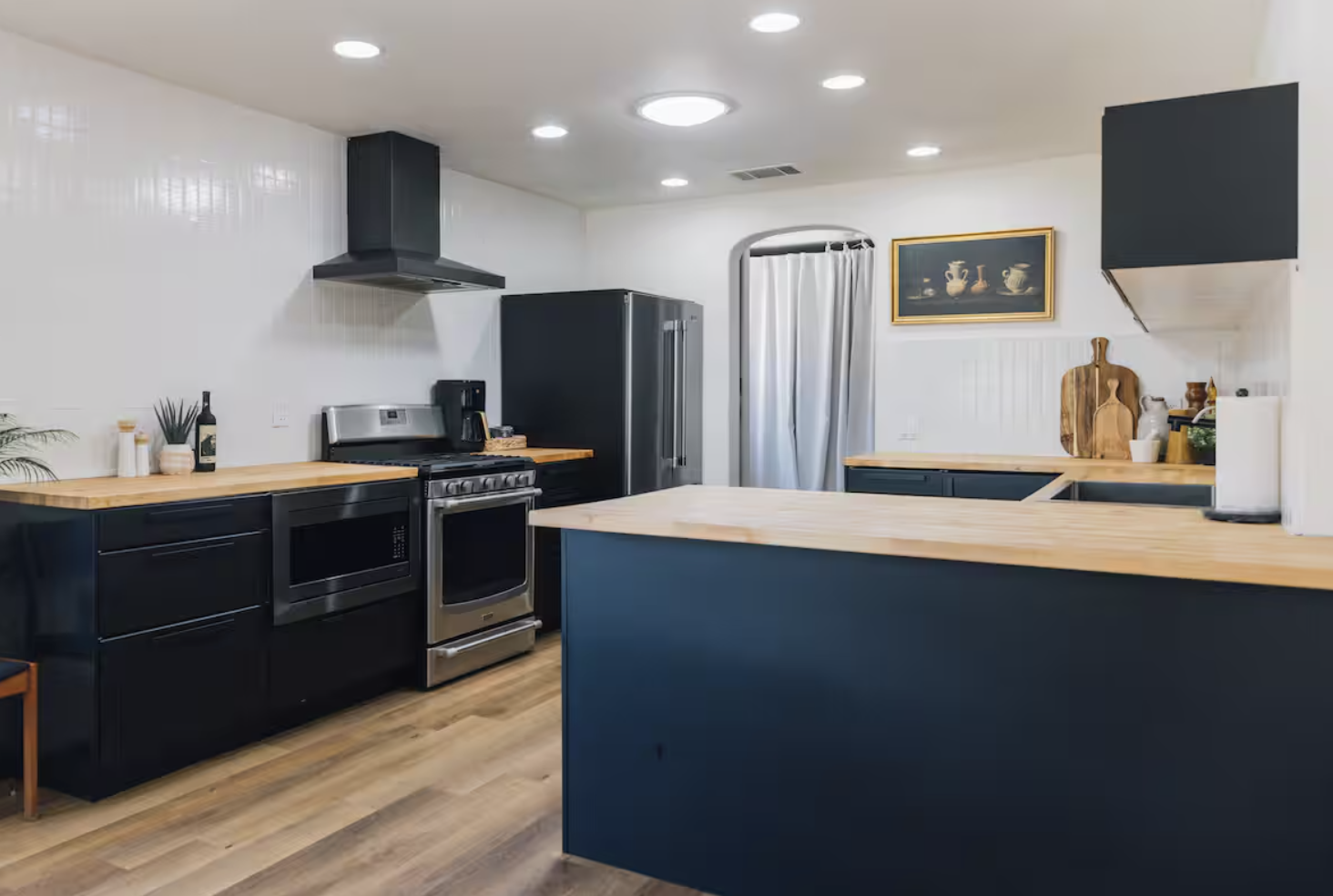 Modern kitchen with black cabinets and light wood countertops, stainless steel appliances, a refrigerator, stove, microwave, and coffee maker. Decor includes cutting boards, potted plants, and framed artwork. The floor is wood, and a curtain covers a doorway in the background.