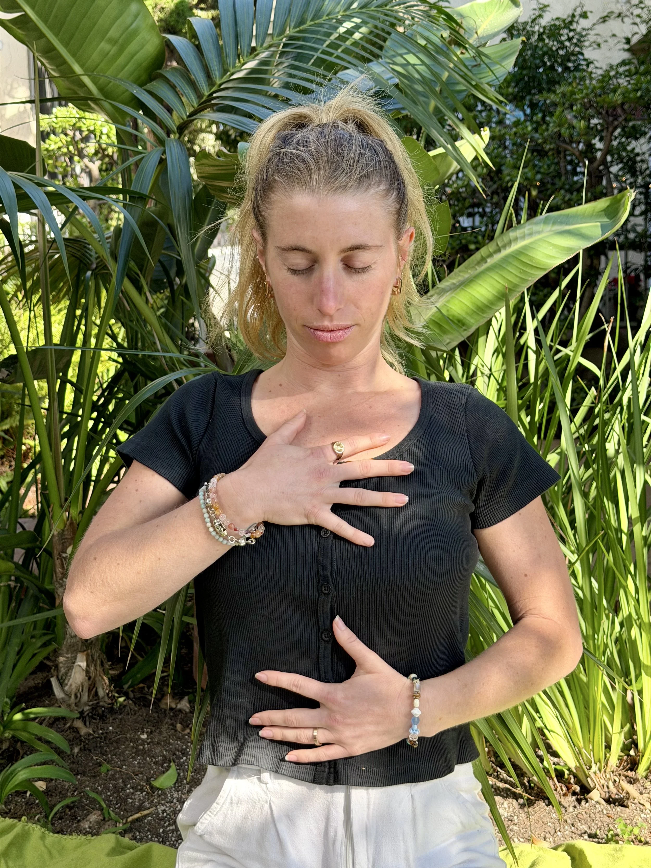 A person with blonde hair and closed eyes standing outdoors among large green tropical plants, wearing a black top and white pants, with one hand on their chest and the other on their stomach, wearing jewelry including bracelets and rings.