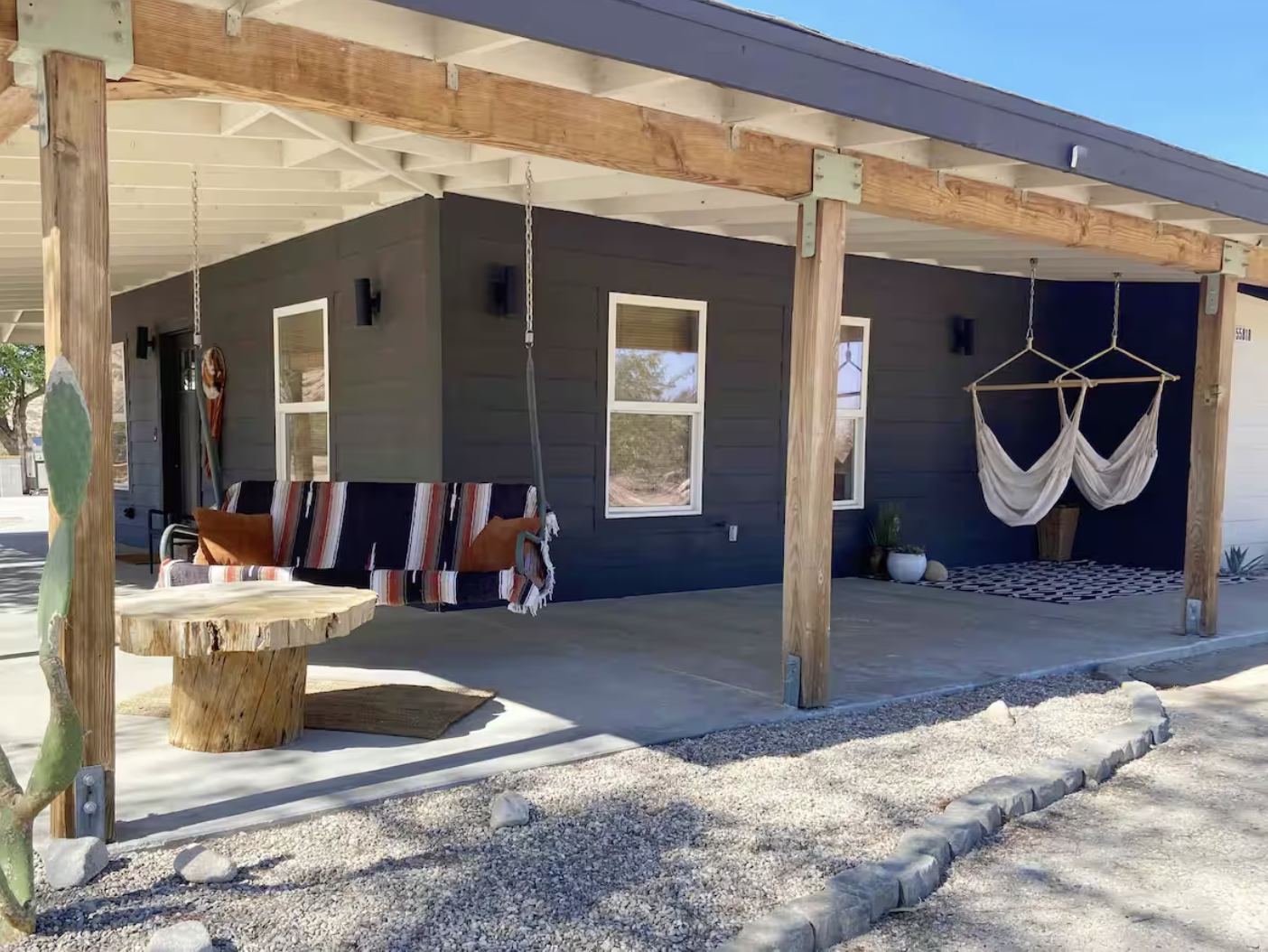 Front porch of a modern house with a swing, hanging hammock, and potted plants, set against a dry, rocky landscape under a clear blue sky.