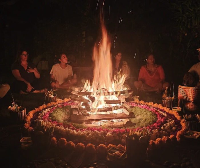 People sitting around a large bonfire at night, with floral decorations surrounding the fire.