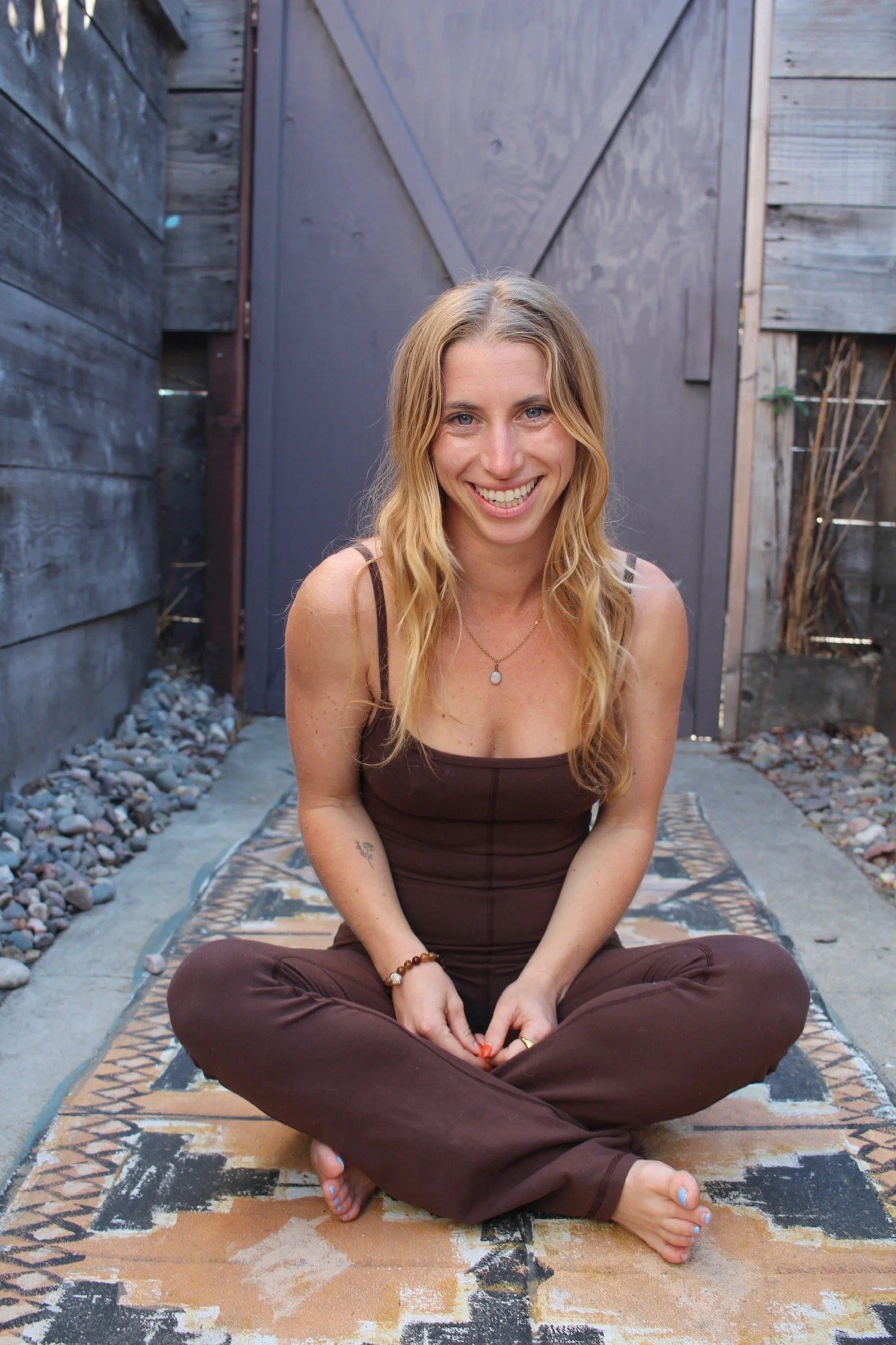 A smiling woman with blonde hair, wearing a brown tank top and brown pants, sitting cross-legged on a patterned outdoor rug, with a background of wooden fence and shed.