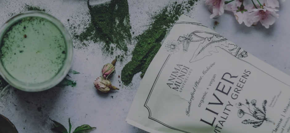 A green smoothie in a glass, green powder spilled on a surface, dried pink roses, and a packet of organic vegan alkaline vitality greens powder from Anima Mundi.