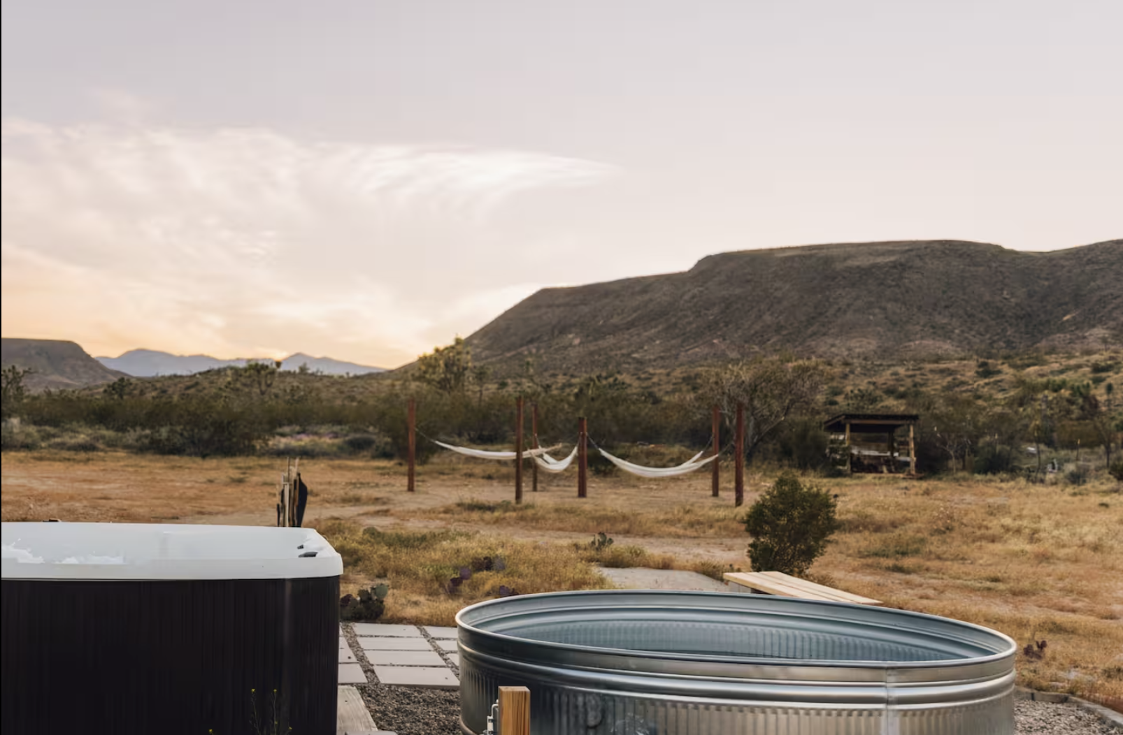 A backyard scene at sunset with hot tubs in the foreground, a hammock tied between wooden posts, and desert mountains in the background.