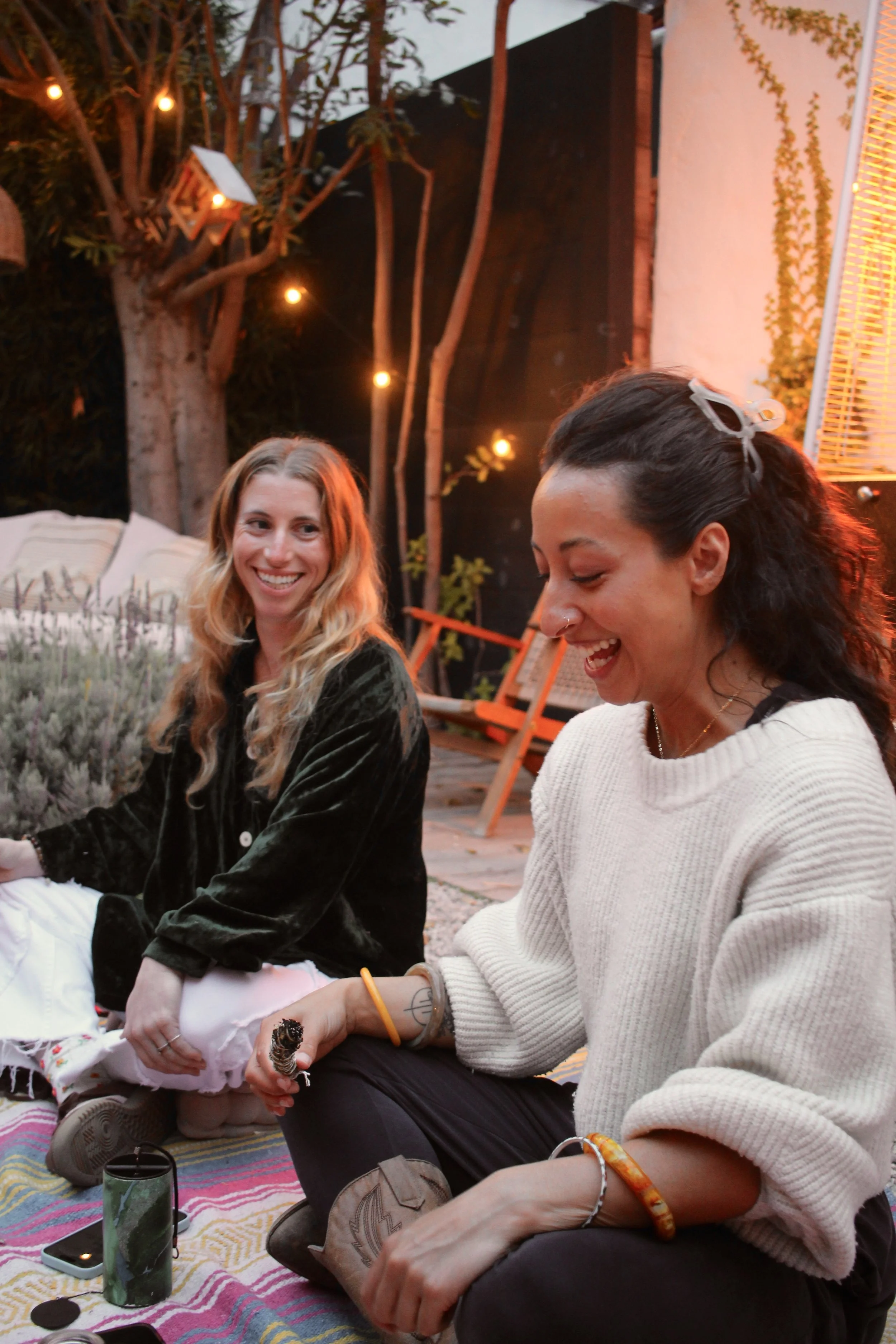 Two women smiling and laughing while sitting on a colorful blanket outdoors during the evening, with string lights and trees in the background.