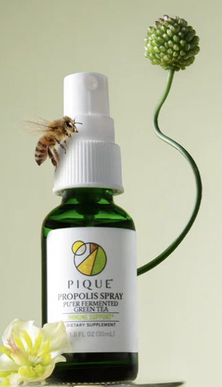 A green bottle of Pique Propolis Spray with a bee on its side, a curly plant stem, a flowering plant, and a flower in the foreground.