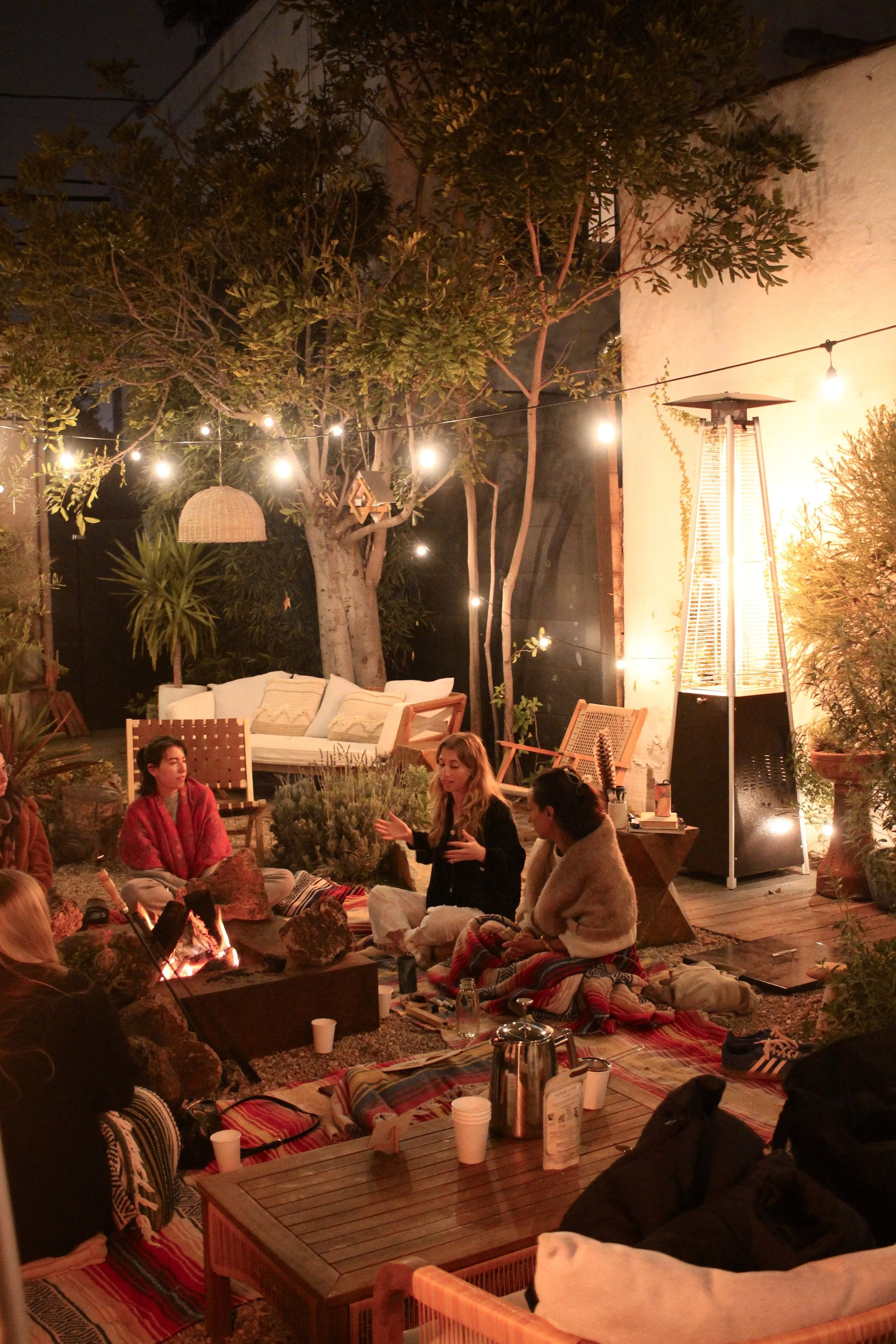 People gather around a fire pit in an outdoor setting with string lights, trees, and patio furniture during evening or night.
