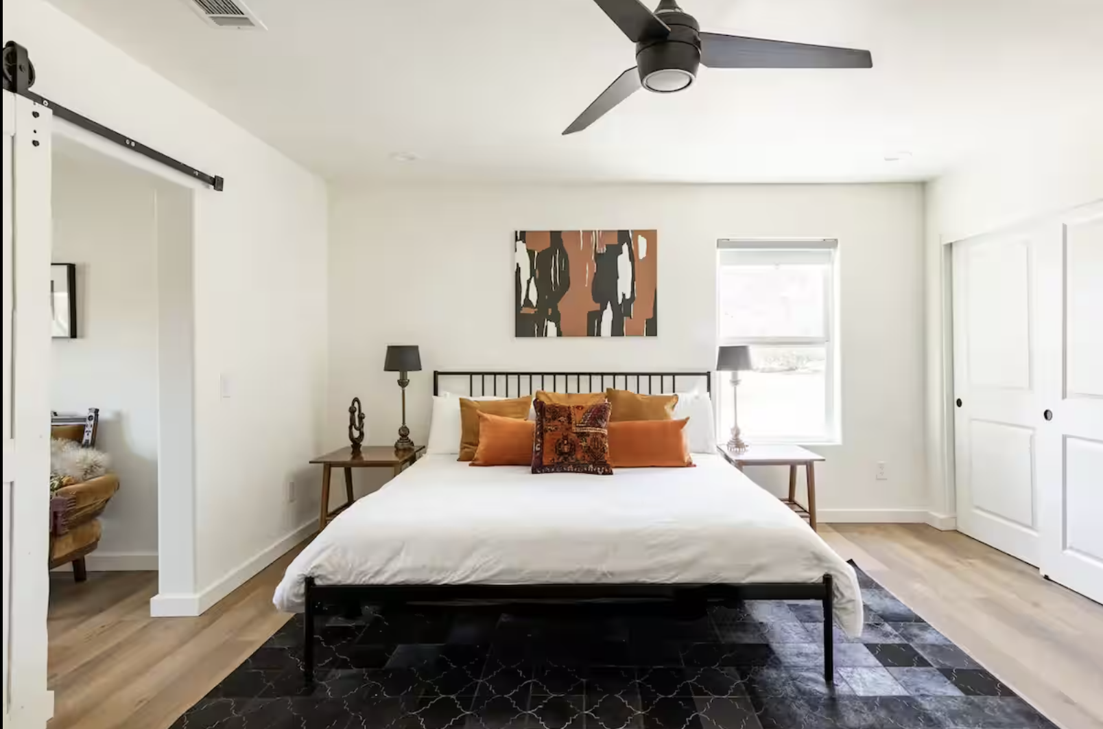 A bedroom with a black metal bed frame, white bedding, and multiple orange and patterned pillows. Two bedside tables with lamps, a window with blinds, and modern artwork above the bed. Light-colored wooden floors and a black accent rug under the bed. A ceiling fan with three blades.