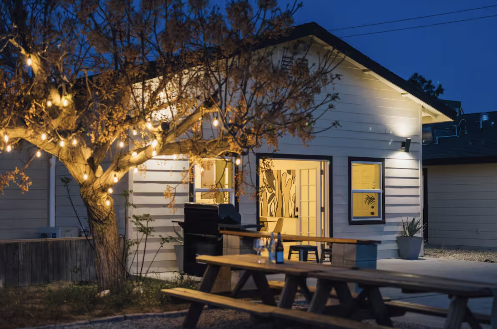 A cozy backyard scene at dusk with a house, lit windows, a deck, a picnic table with drinks, and a tree with string lights.