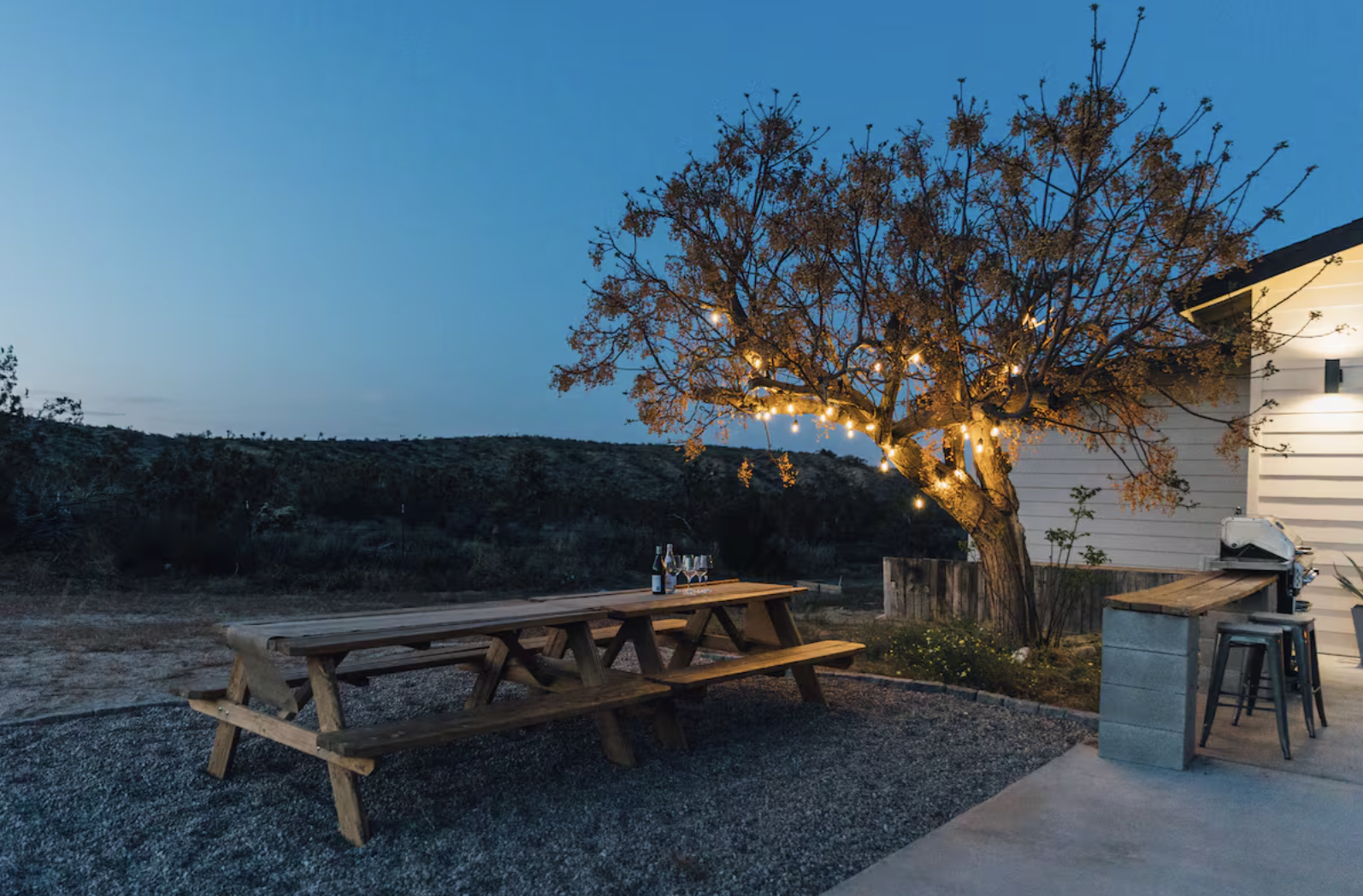 Outdoor patio scene at dusk with a wooden picnic table holding bottles and glasses, a lit tree decorated with string lights, a house with a bar-height counter and barstool, and a barbecue grill.