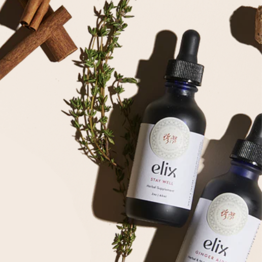 Two black dropper bottles labeled 'elix Stay Well' and 'elix Ginger' on a white surface with cinnamon sticks, a sprig of thyme, and wooden pieces nearby.