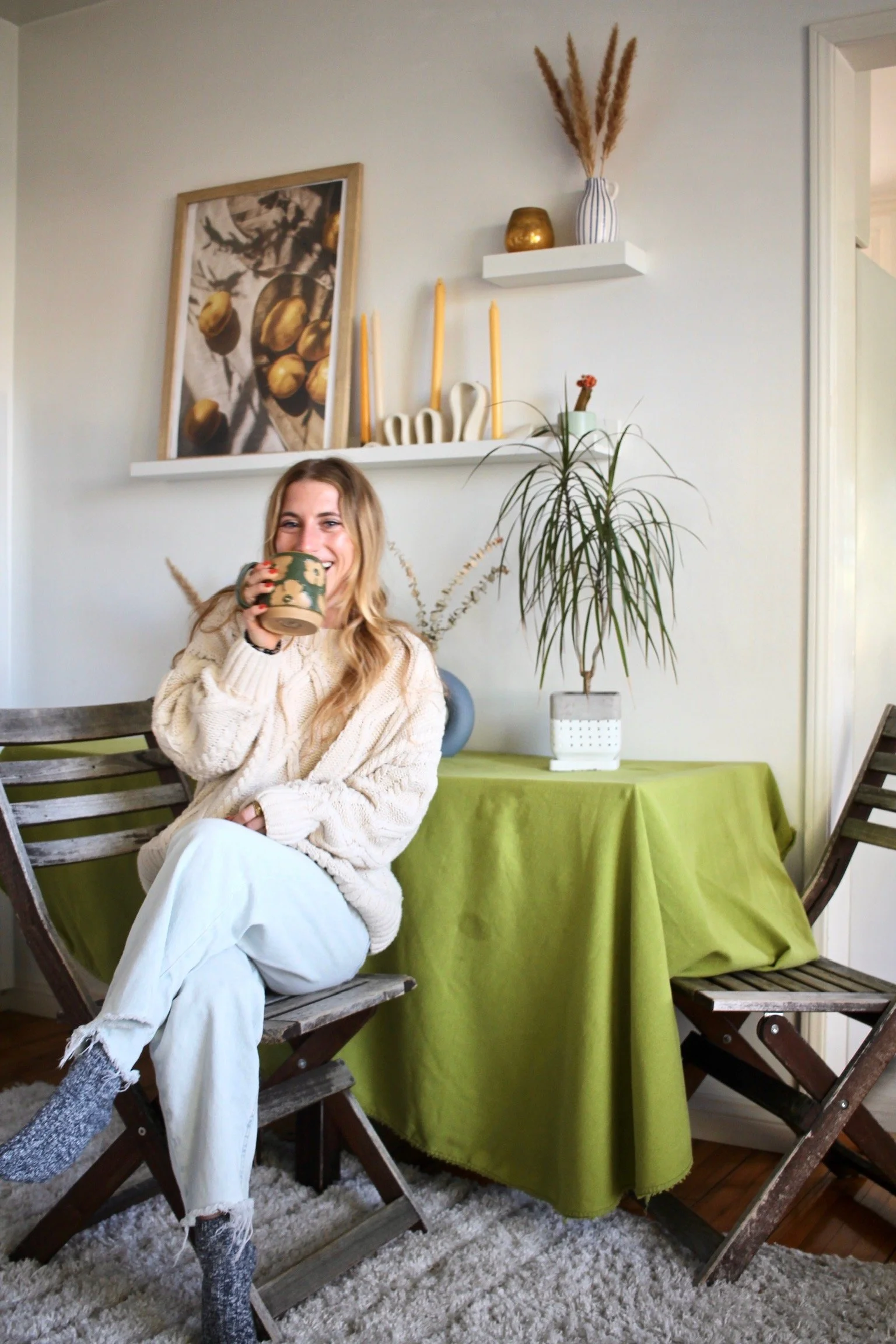 A woman sitting on a wooden chair holding a mug with a floral pattern. She has long blonde hair and is smiling. She is wearing a beige knitted sweater, white ripped jeans, and gray socks. Behind her, there is a table with a green tablecloth, a large potted plant, and decorative items on a white shelf, including a framed picture of pears, candles, and vases.