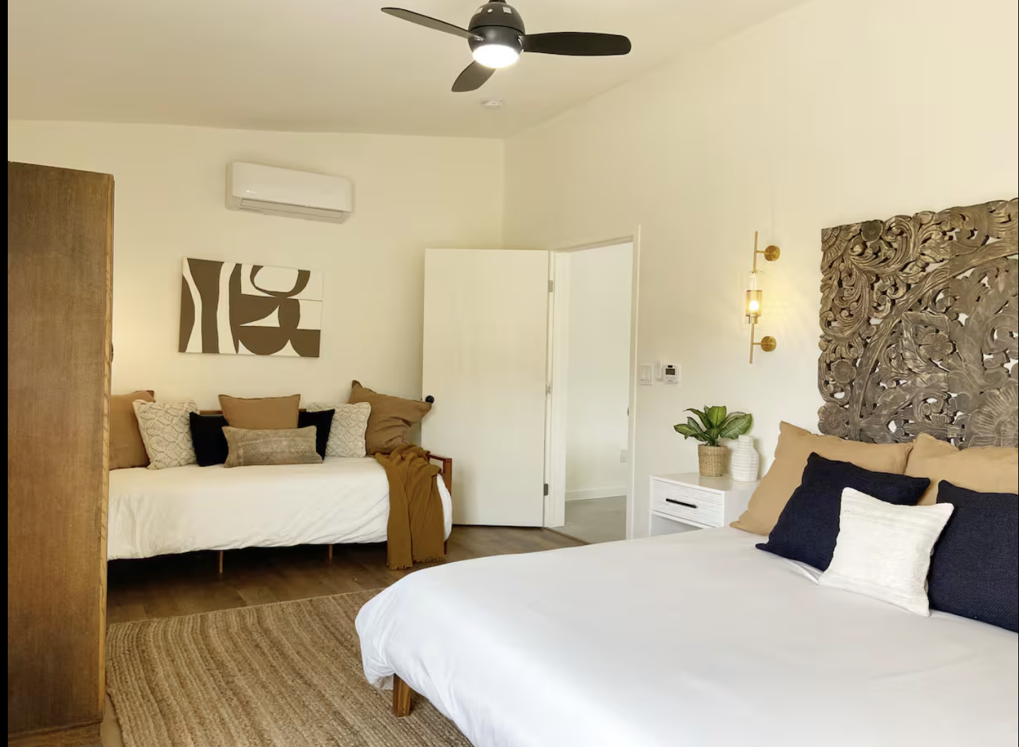 A bedroom with two beds, one with a large carved wooden headboard, and another with a wooden bench and various pillows. The room has white walls, a ceiling fan, wall-mounted light fixtures, and a potted plant on a white nightstand.