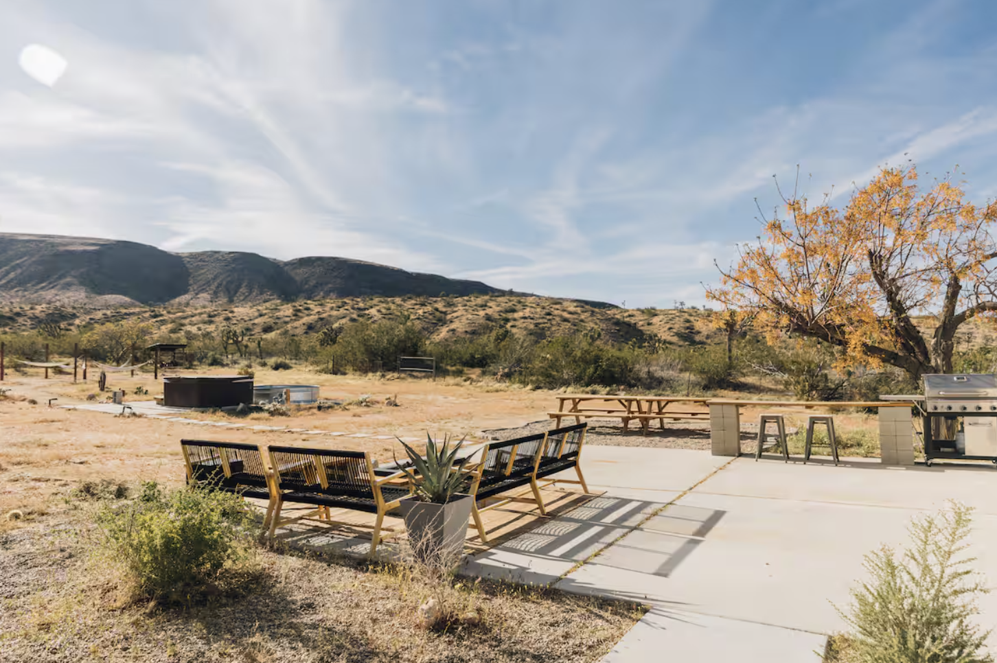 Outdoor patio area with benches, potted plants, a grill, and picnic tables in a dry, desert-like landscape with mountains and a tree with orange leaves under a partly cloudy sky.
