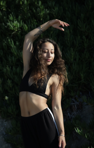 Young woman with curly hair standing outdoors in front of green foliage, wearing a black sports bra and black pants, with her eyes closed and one arm raised above her head.