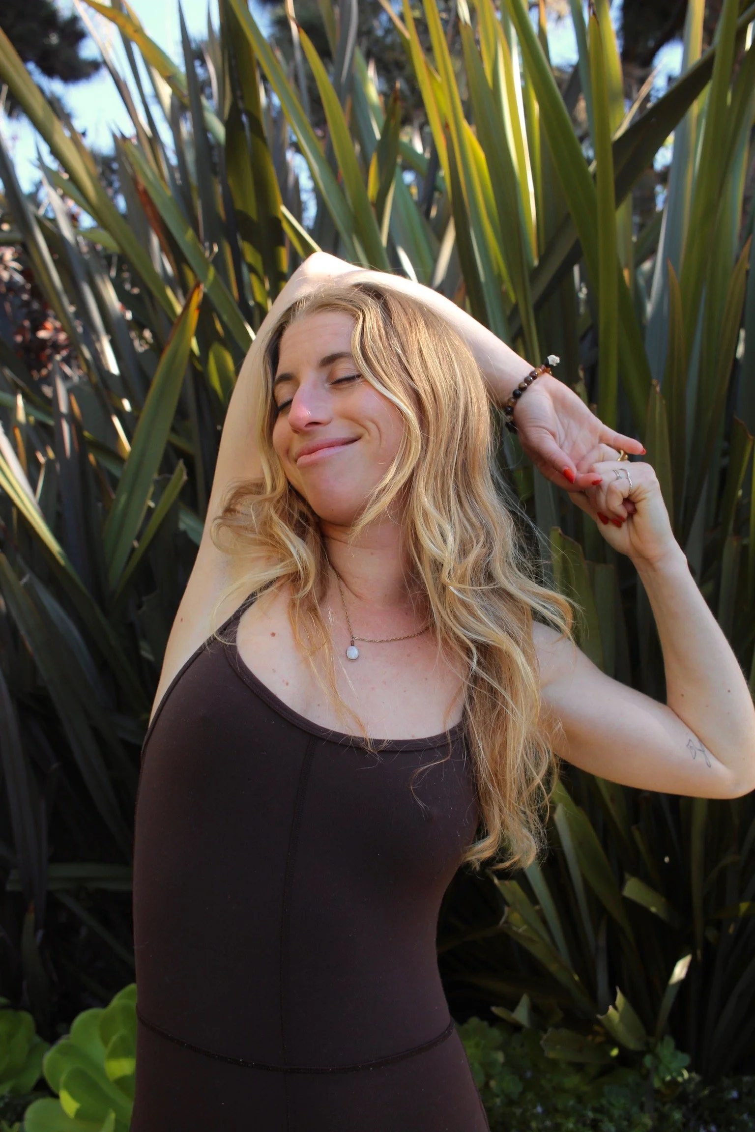 A woman with long wavy blonde hair, closed eyes, and a gentle smile, is posing outdoors among tall green plants with large leaves, with her arms behind her head and wearing a dark sleeveless top and a beaded bracelet.