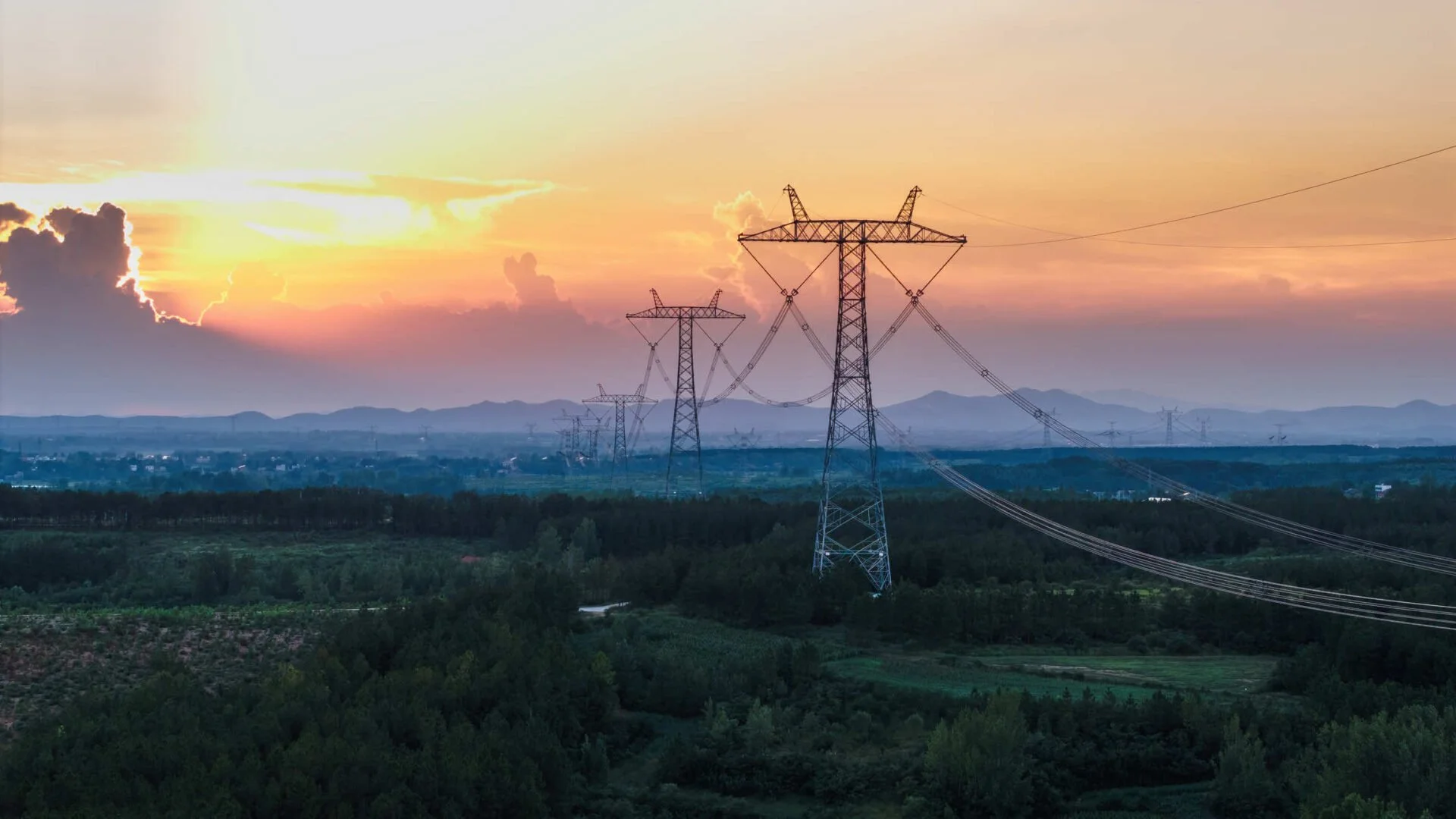 Sunset over a landscape with power lines and green fields, mountains in the background.