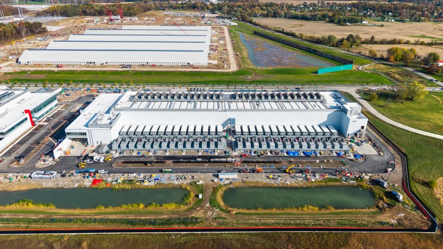 Aerial view of a large manufacturing or distribution facility under construction, with multiple loading docks, construction vehicles, and workers, surrounded by water and open land.