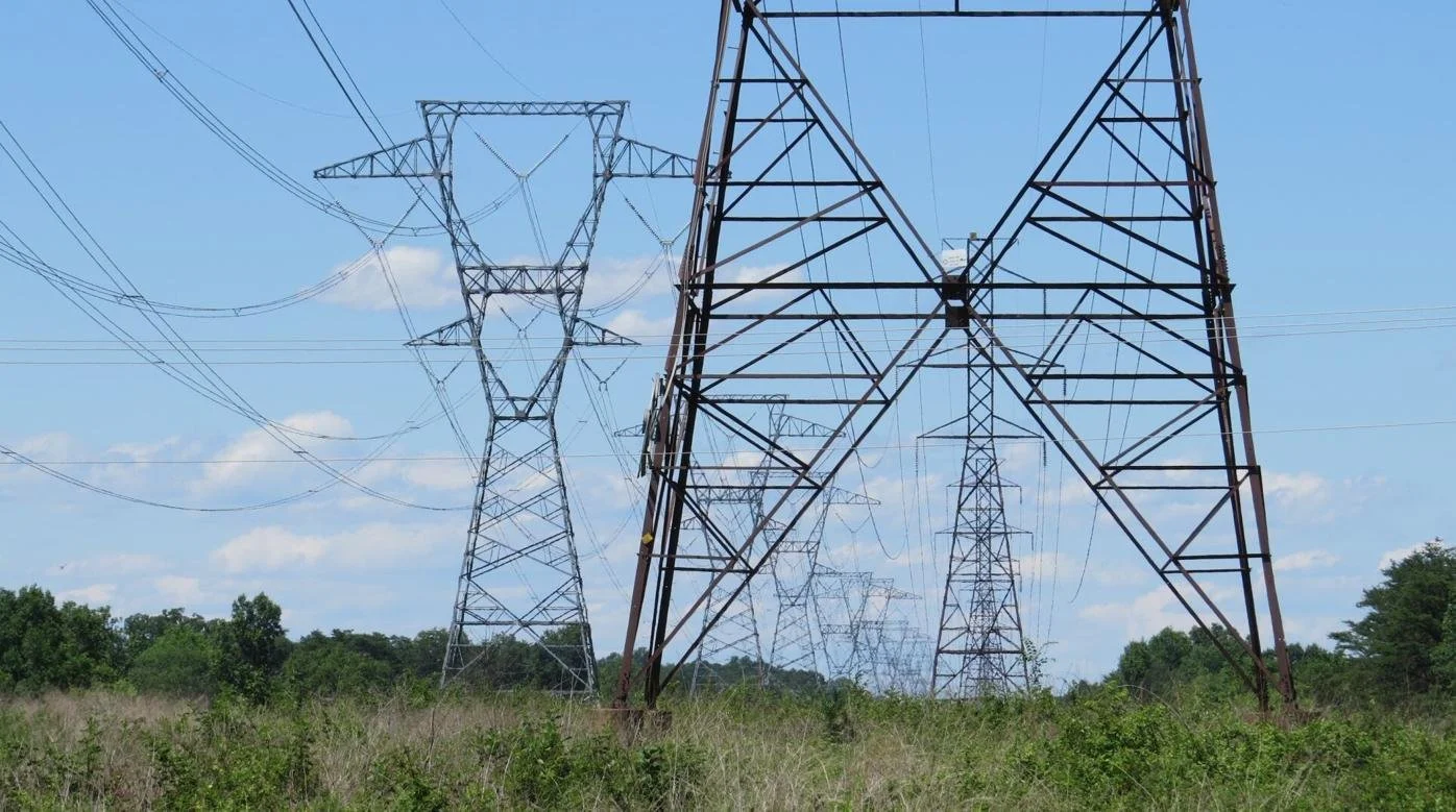 Multiple metal electric transmission towers and power lines stretch across a grassy field with trees in the background under a partly cloudy sky.
