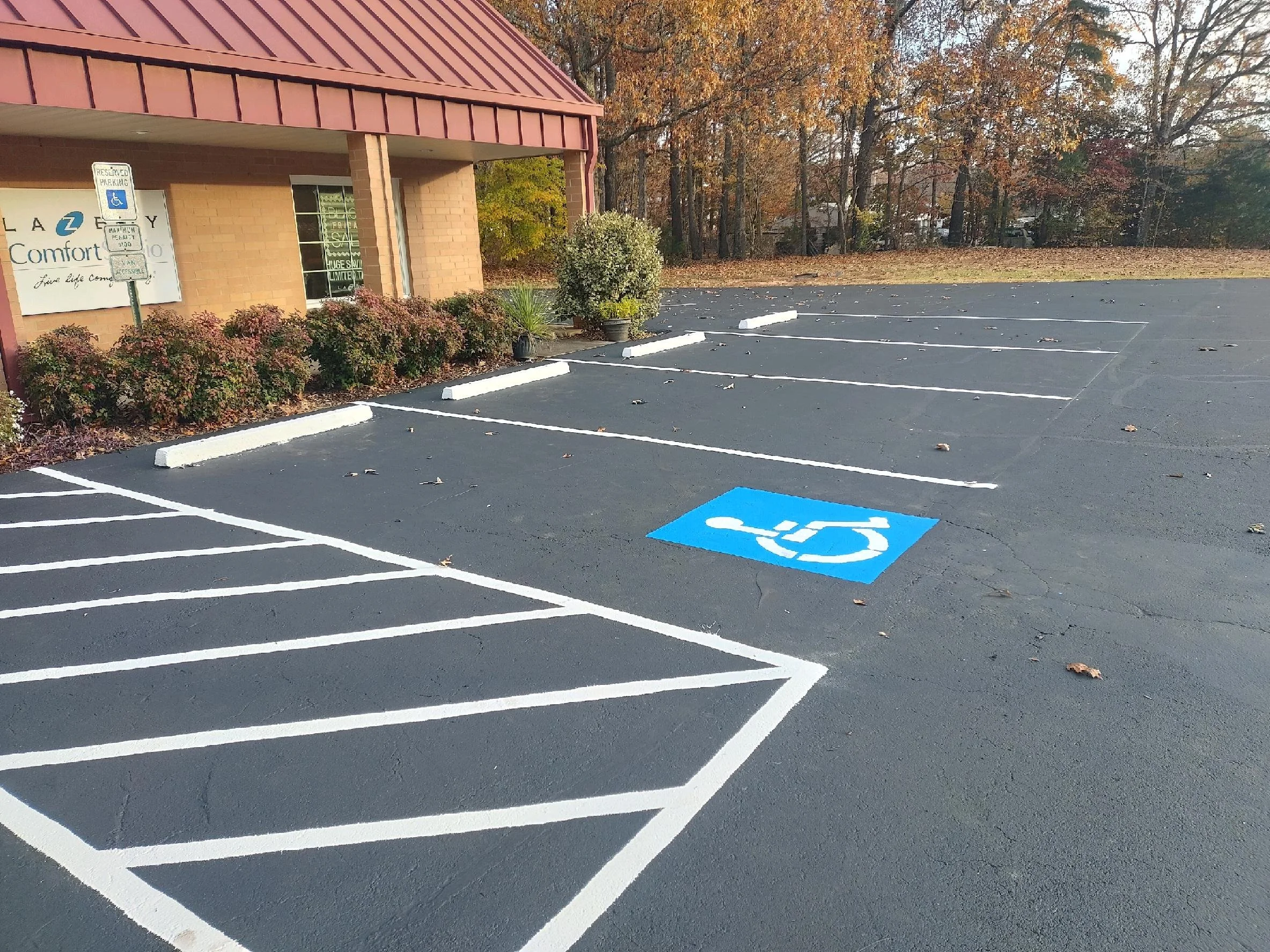 Empty parking lot with a designated handicapped parking space marked in blue, and other parking spots outlined in white. A building with a brick facade and red roof is visible on the left, with a reserved parking sign near the building. Autumn trees with colorful leaves are in the background.