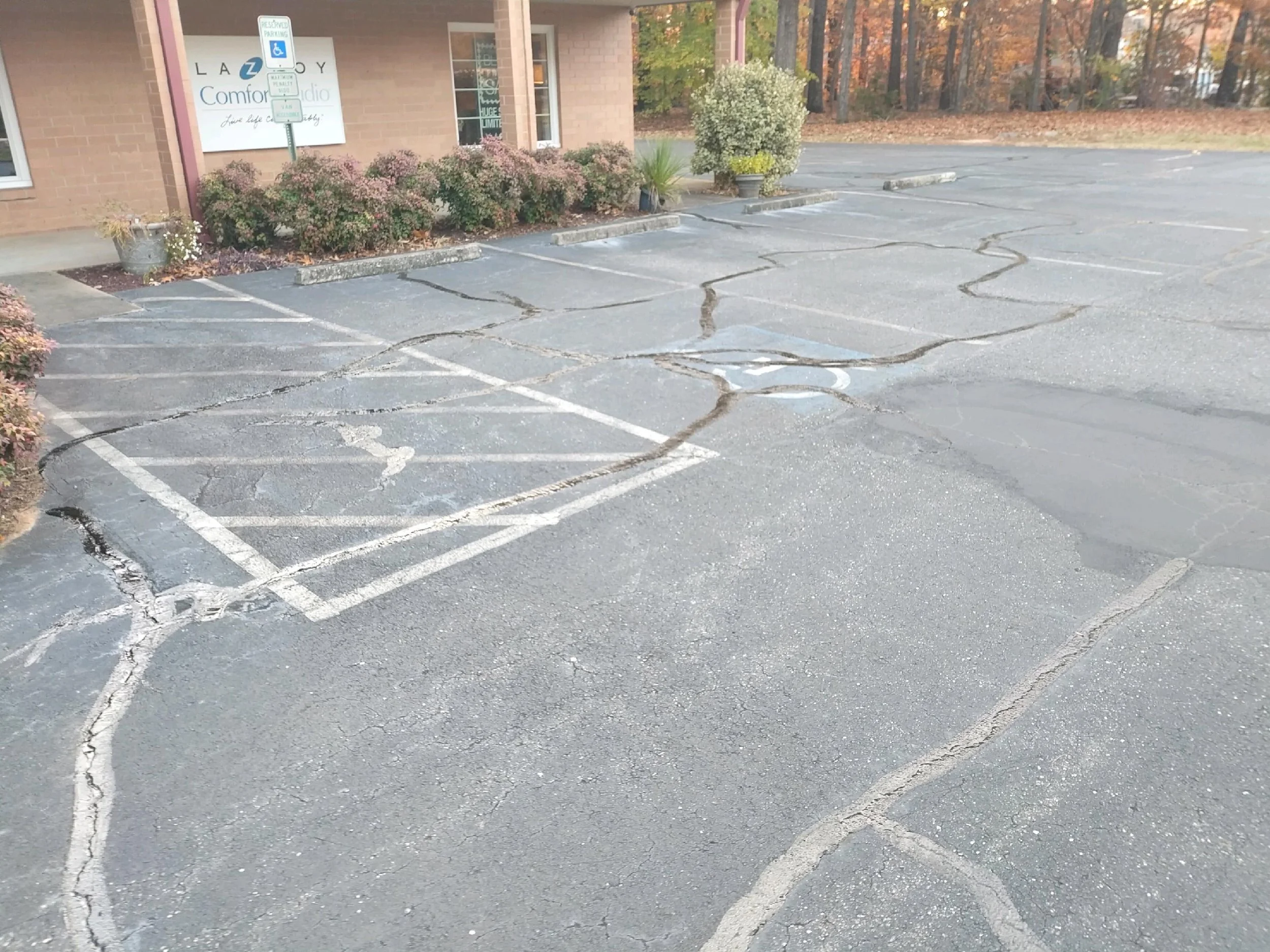 An empty parking lot with cracked asphalt, surrounding bushes, a brick building, and fall trees in the background. There is a sign indicating reserved parking for handicapped.