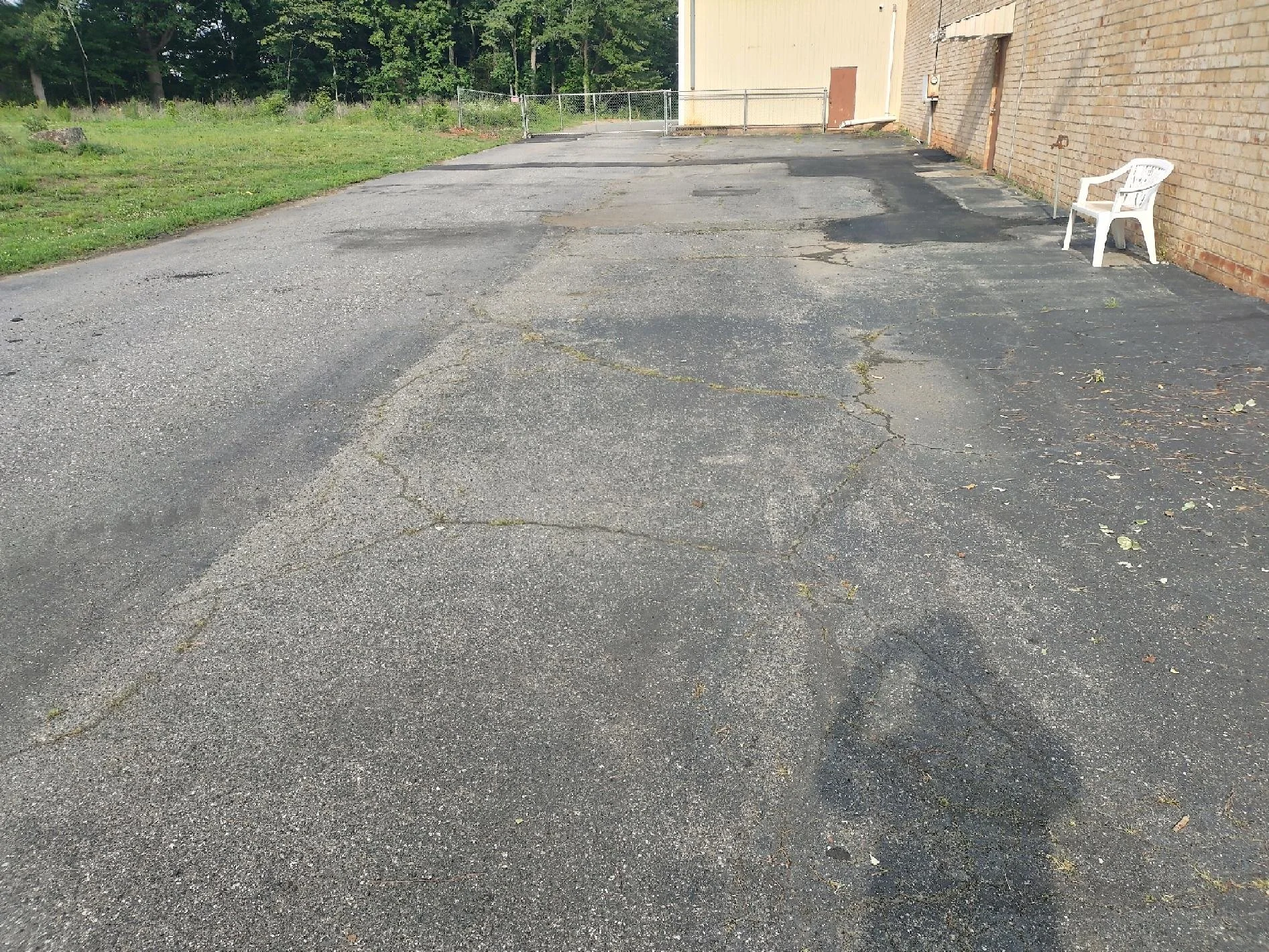 A cracked and weathered asphalt parking lot next to a brick building with a white plastic chair on the right side and a chain-link fence at the far end, with trees in the background.
