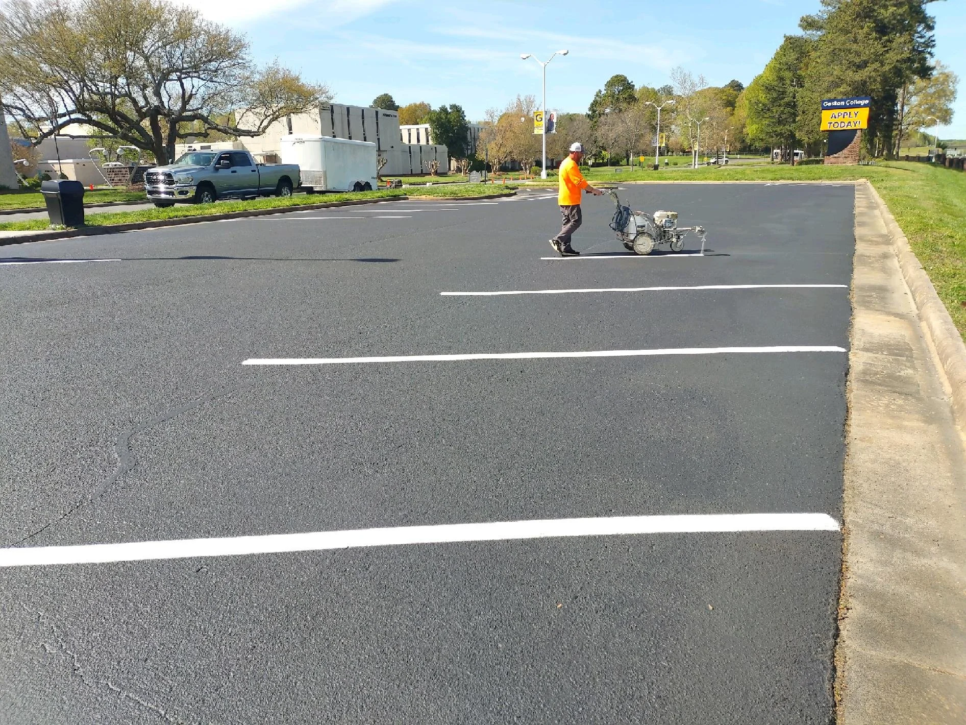 A person in an orange shirt walks beside a street paving machine in a mostly empty parking lot with marked parking spaces, a tree, and a sign that reads "Gaston College APPLY TODAY!" in the background.