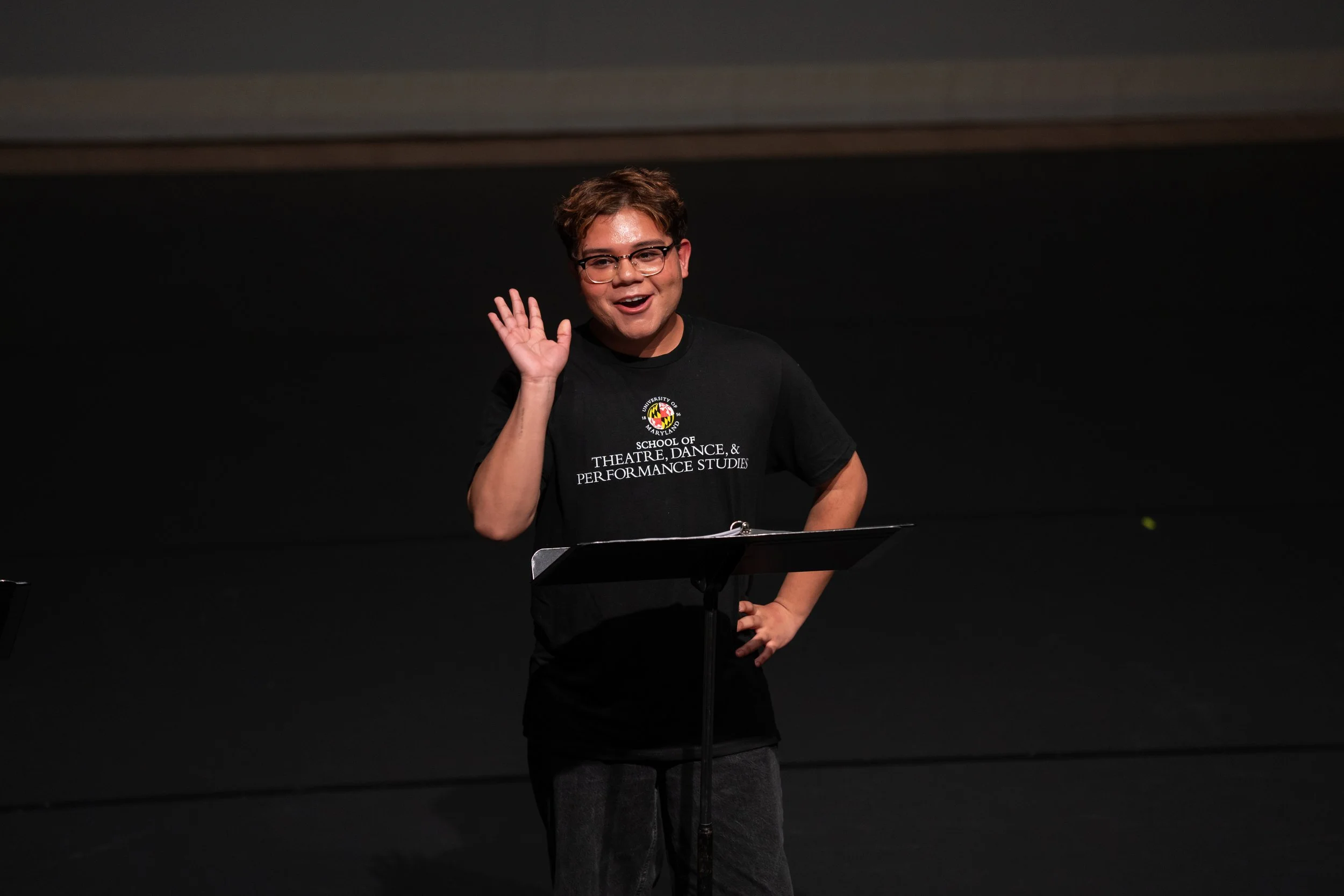 A young man with glasses waving and smiling on stage, wearing a black t-shirt with white text and a logo, standing next to a music stand.