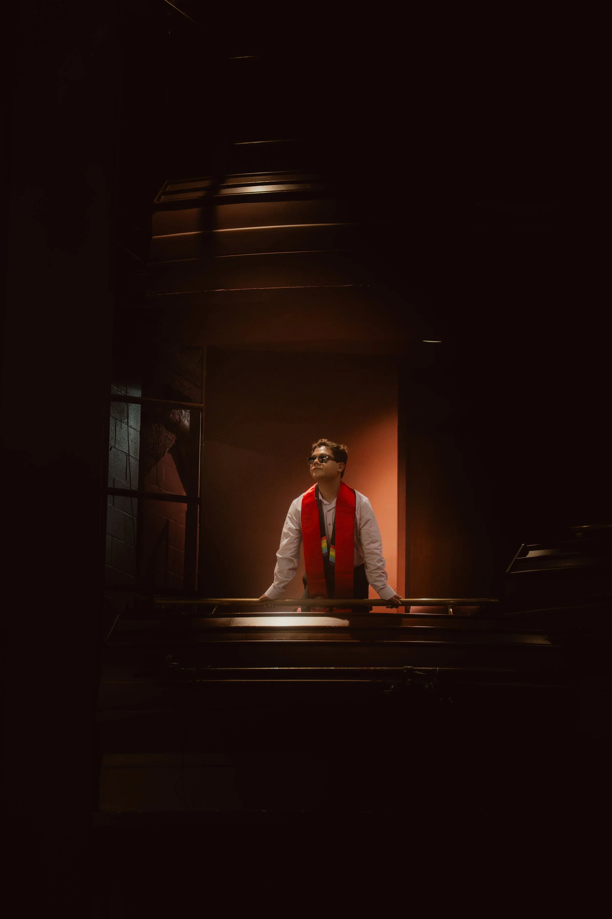 A man in a white shirt, black tie, and wearing glasses, standing behind a piano with a red stole around his neck, appears to be in a dimly lit stage or performance setting with warm lighting focused on him.