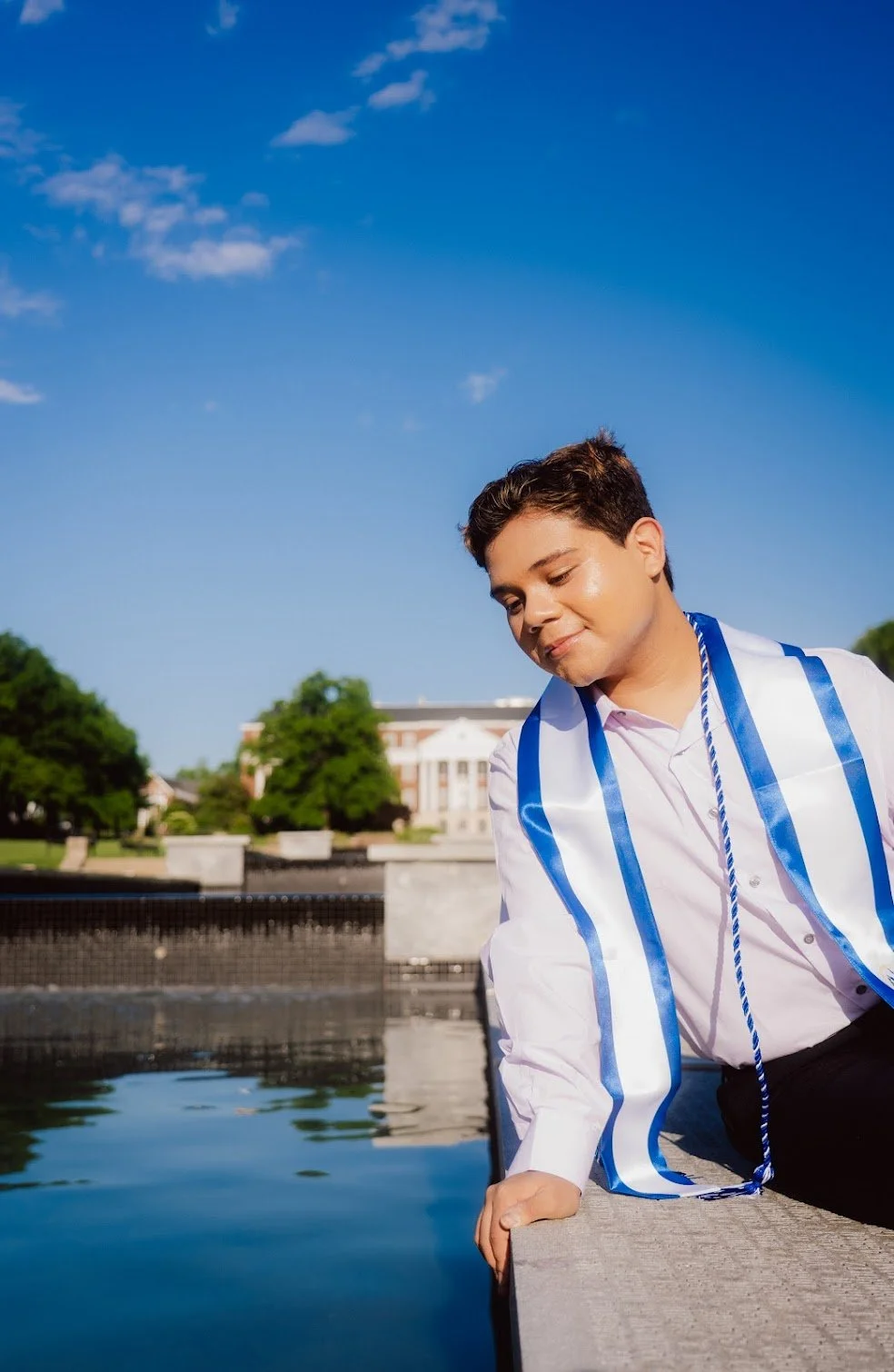 A young man in a white shirt and blue tassel stole leaning on a stone ledge next to a body of water, with a graduation sash around his neck, smiling with eyes closed under a clear blue sky, in front of a university campus with trees and a white building.