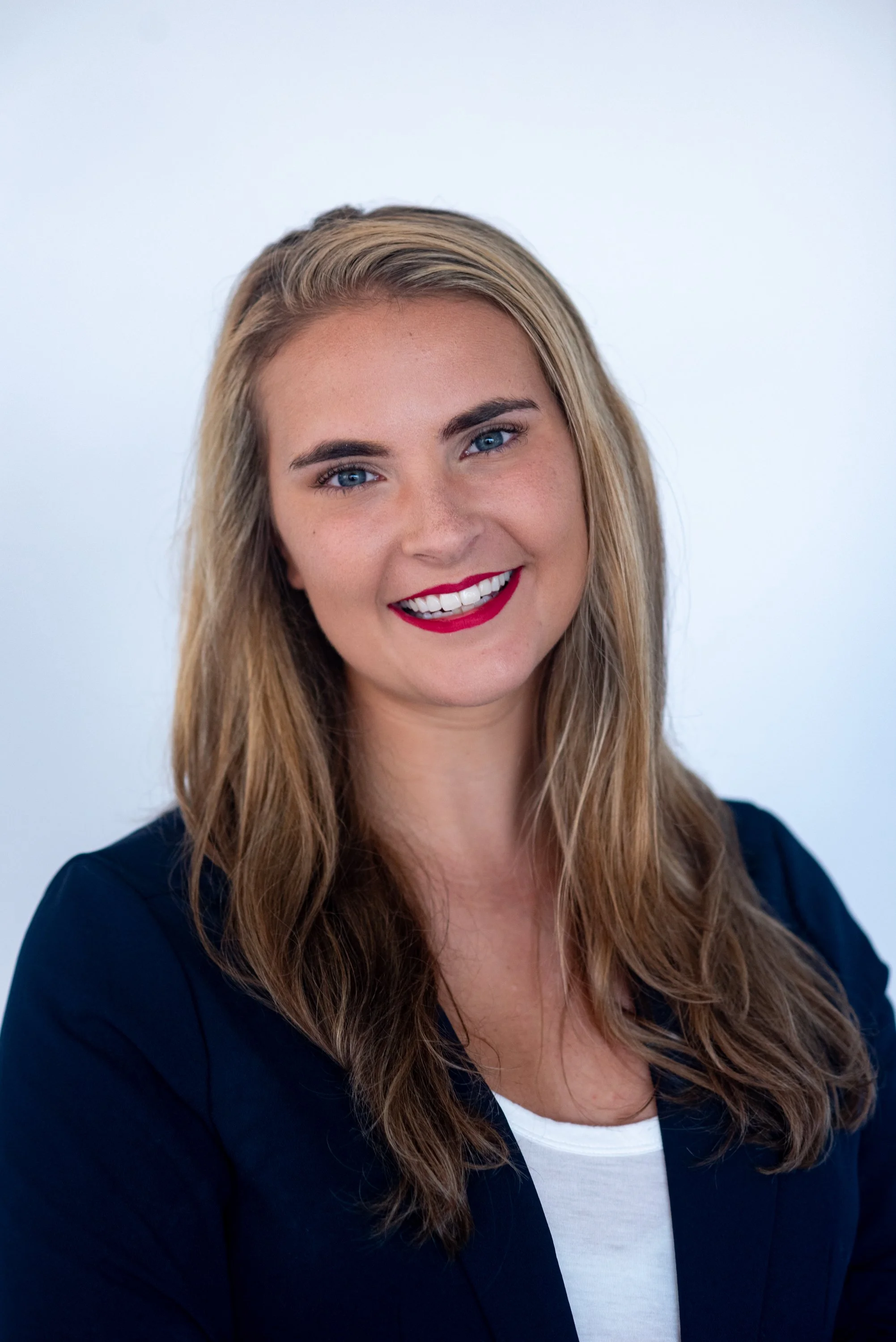 A woman with long wavy blonde hair, light skin, bright blue eyes, wearing a navy blue blazer over a white top, smiling against a plain white background.