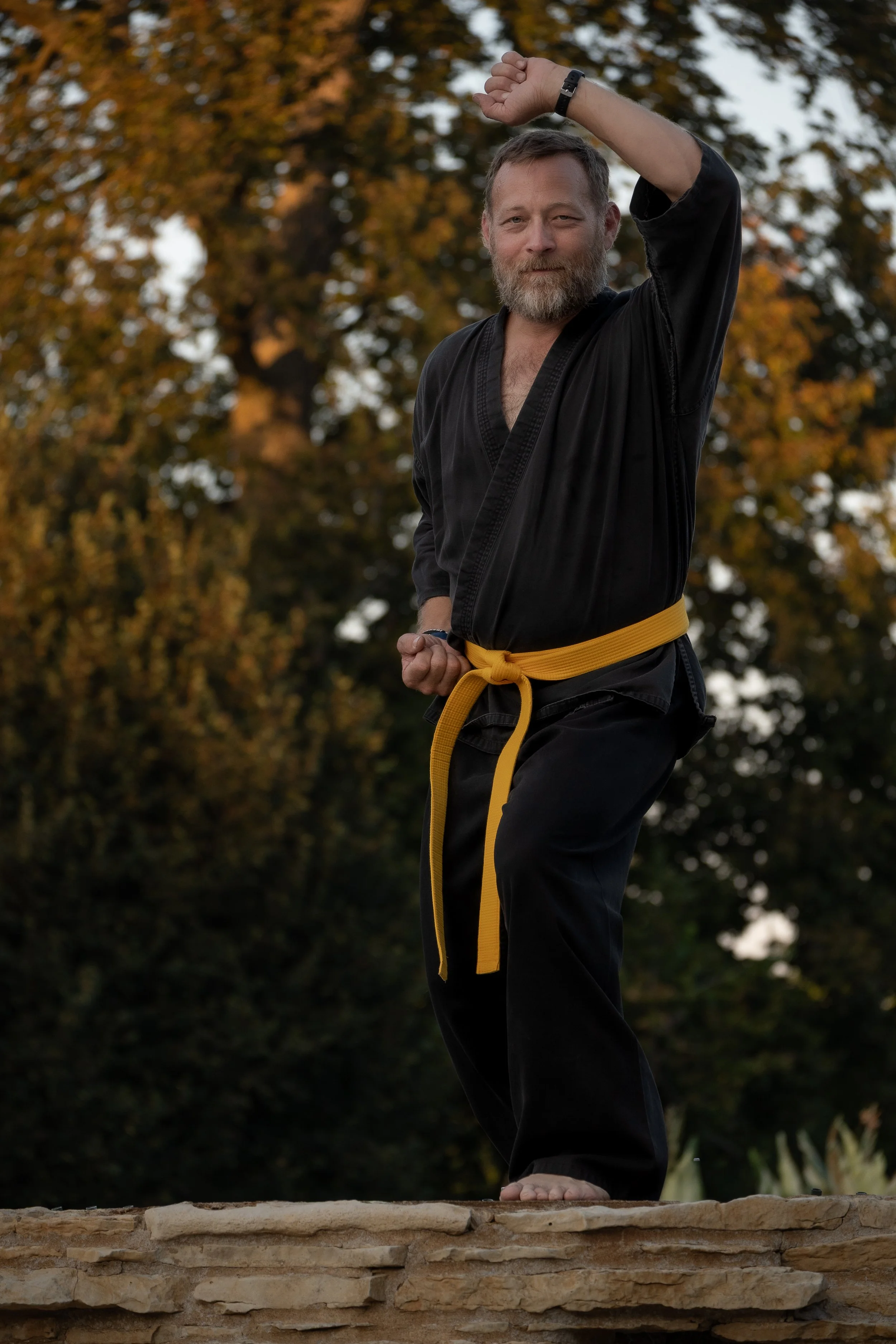 A man in a black martial arts uniform with a yellow belt practicing yoga outdoors on a stone platform, surrounded by autumn trees.
