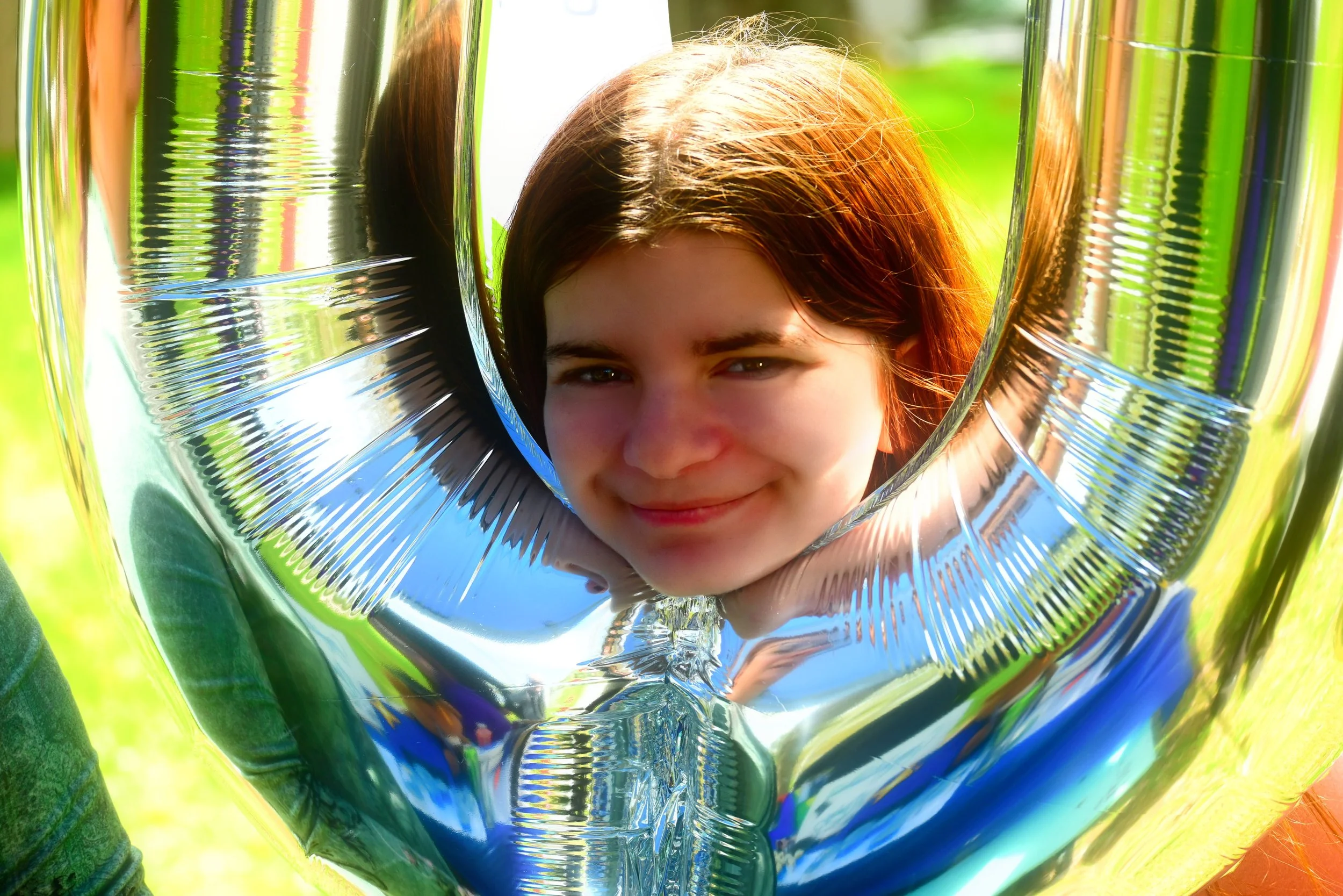 A young girl with brown hair and a smile holding a shiny silver balloon outdoors with green grass in the background.