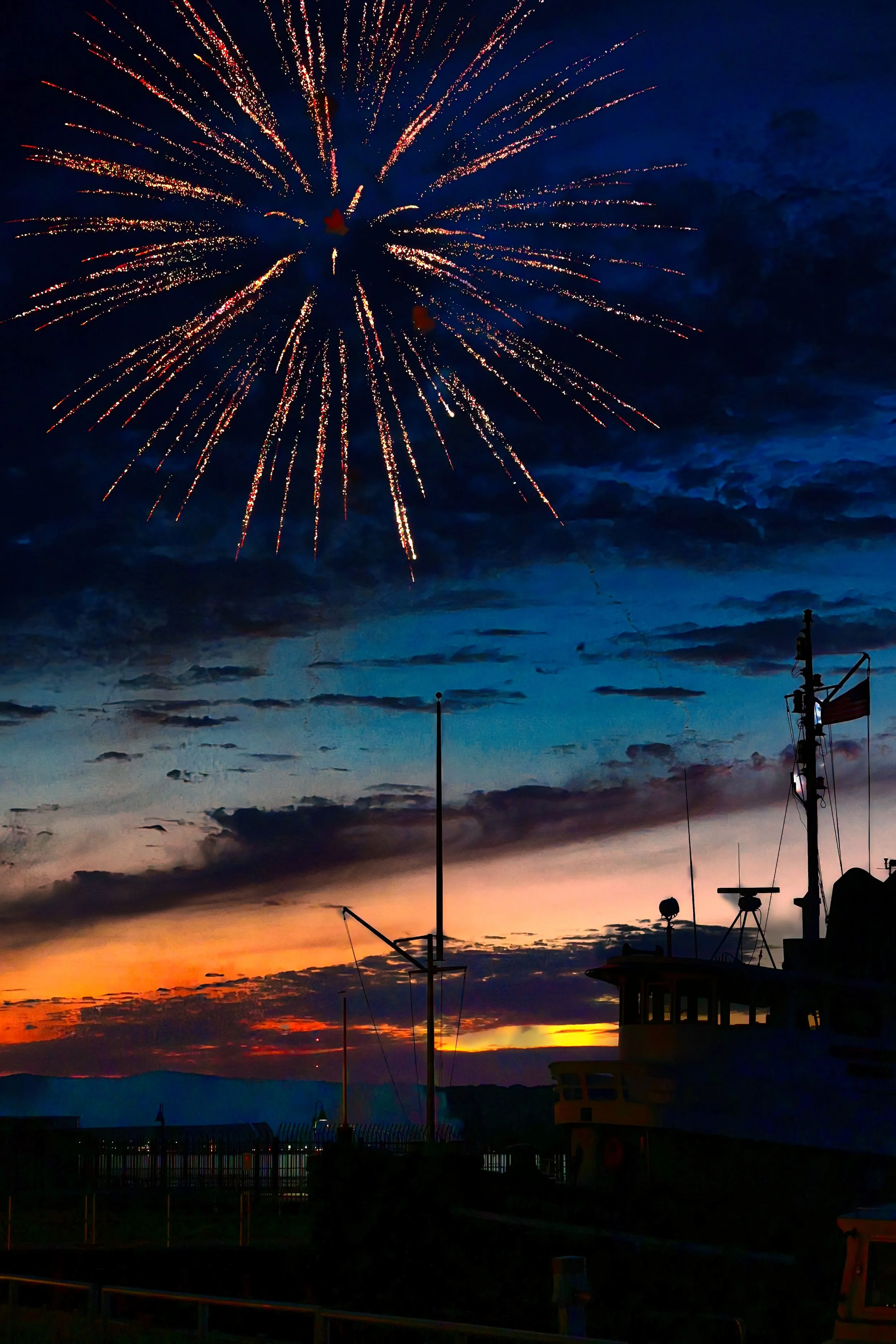 Fireworks exploding in the night sky over a harbor with boats, including a large ship, during sunset with orange and blue colors.