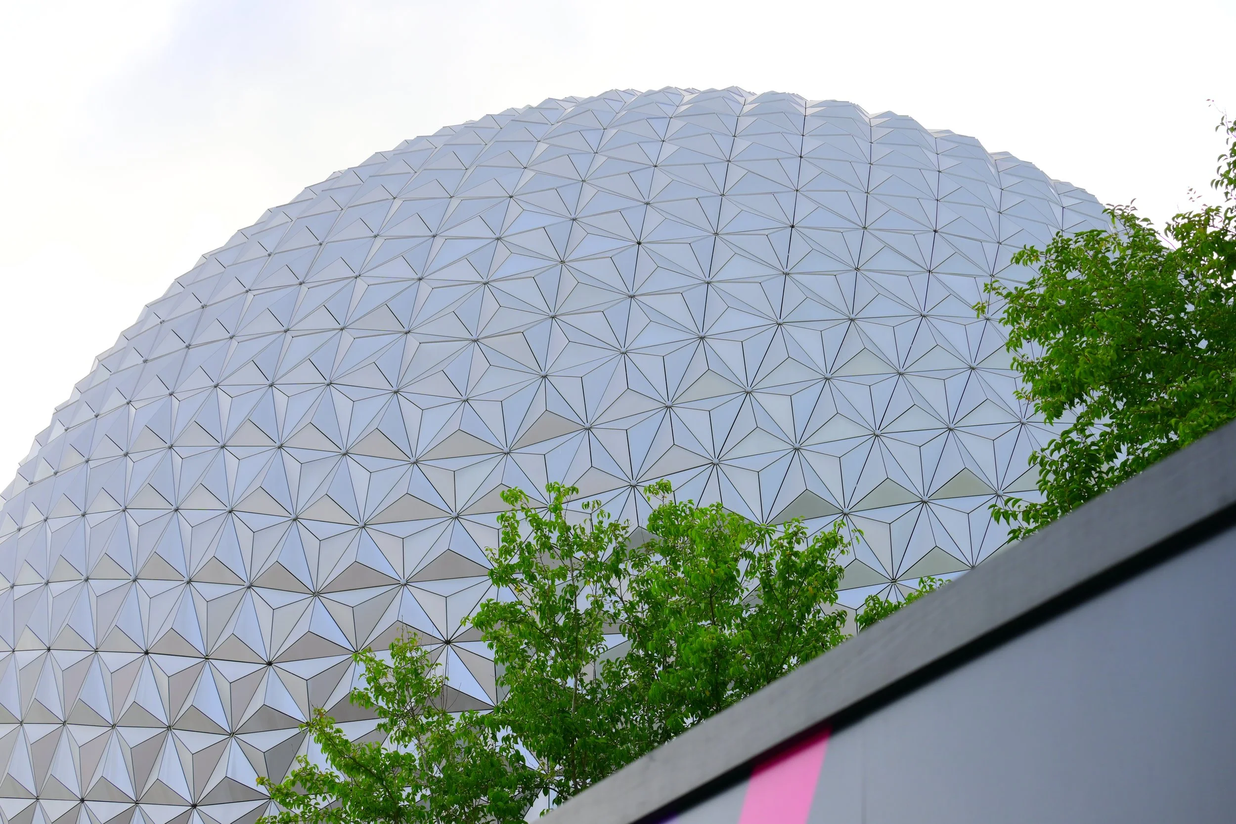 Low-angle view of a geodesic dome structure with interlocking panels, partially obscured by green trees, against a cloudy sky.