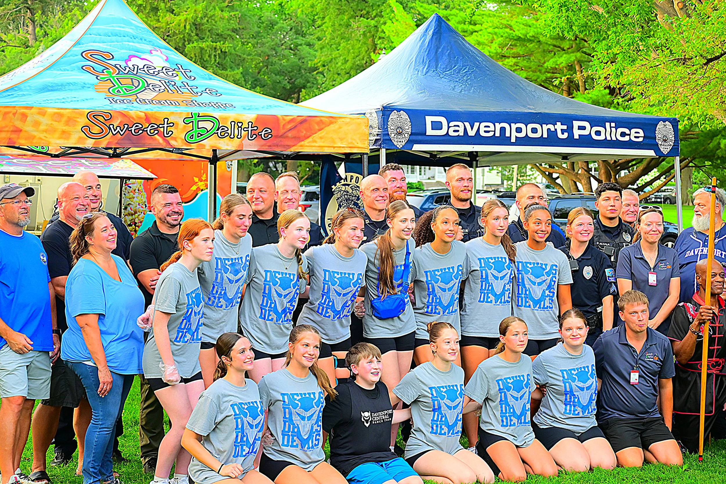 Group of children and adults wearing Davenport Demons shirts, standing together outdoors in front of tents and trees, during a community event.
