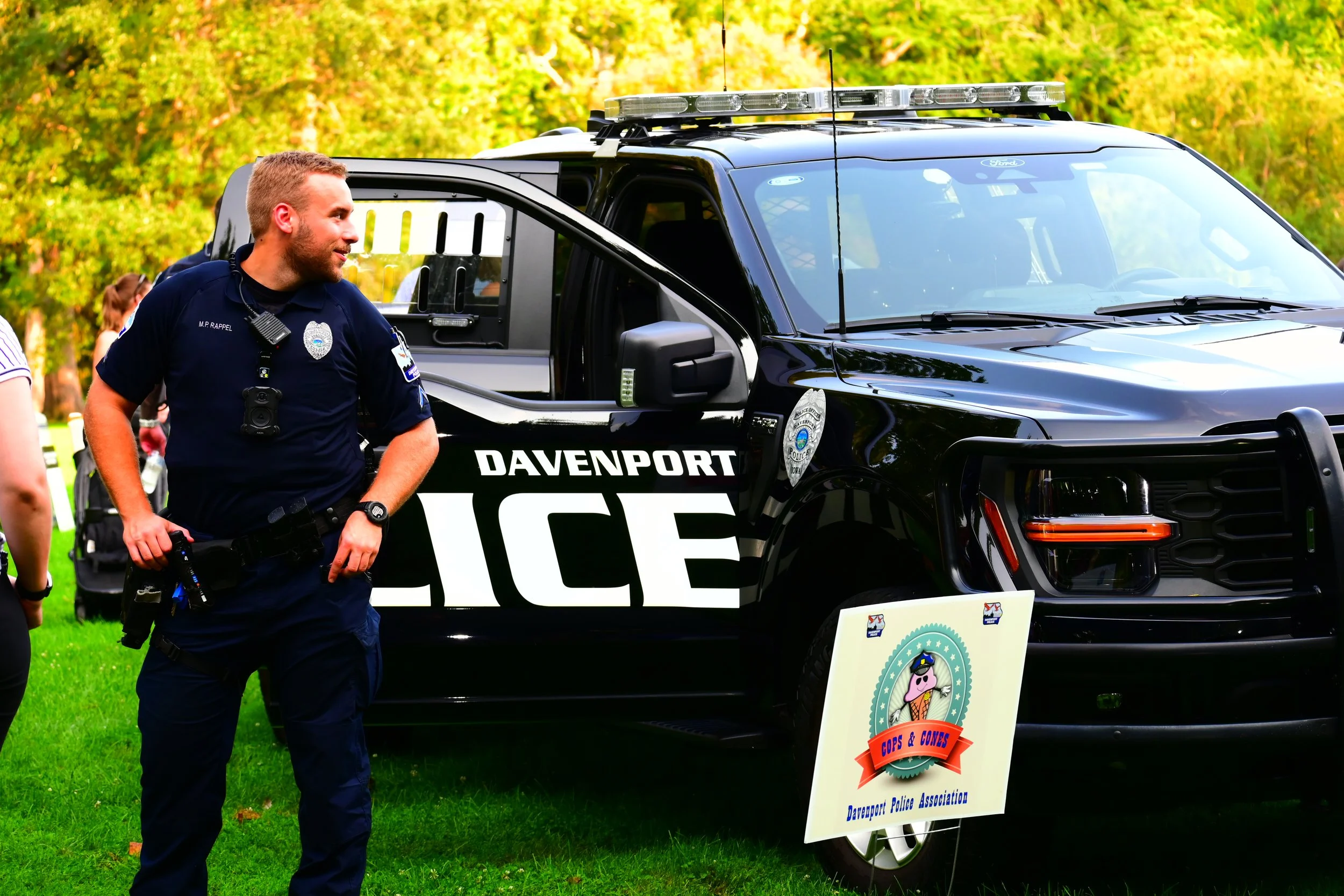 A police officer standing outdoors near a black police vehicle with emergency lights on the roof during a community event. There are trees and a colorful sky in the background.